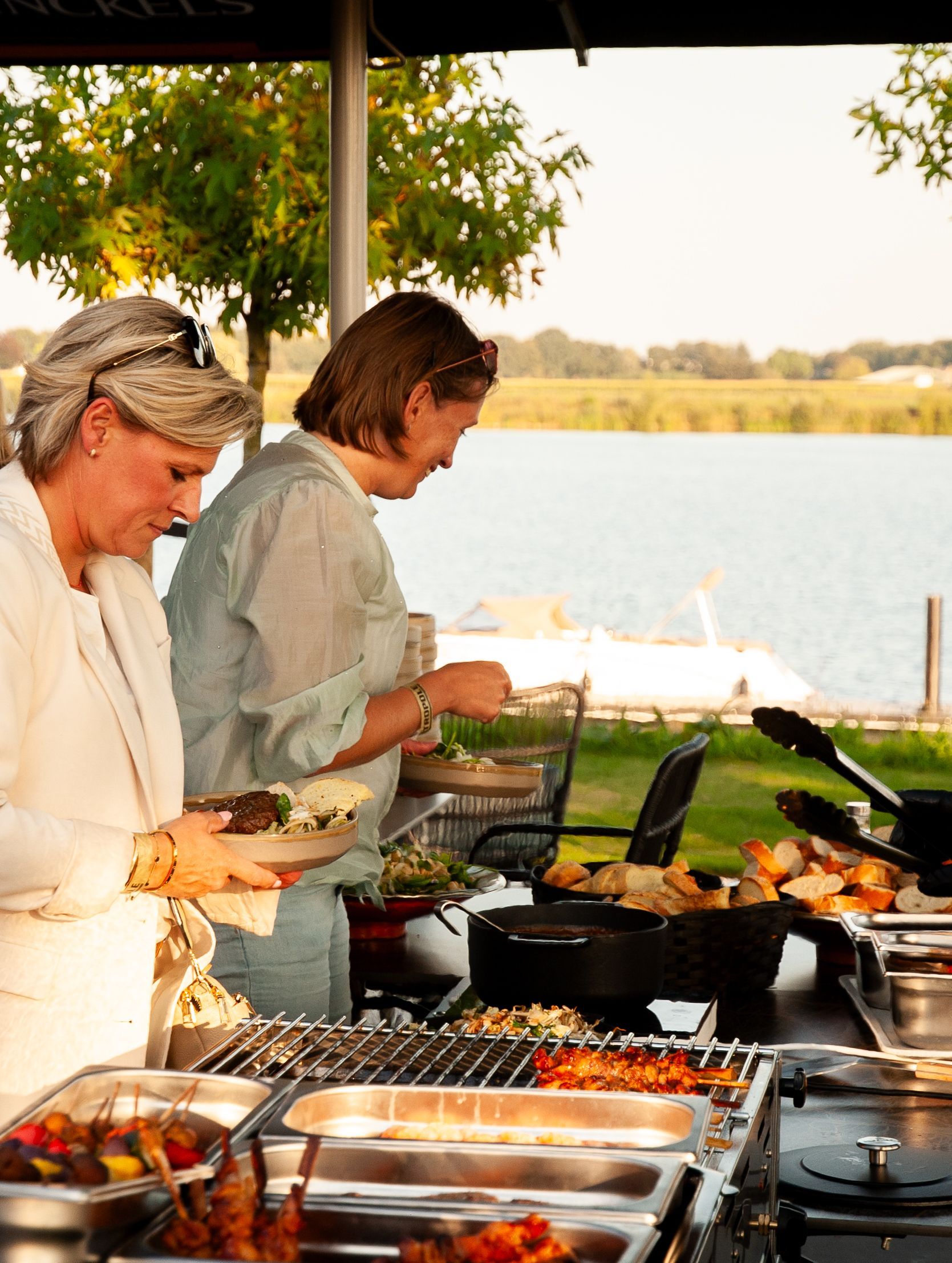 Twee mensen serveren eten bij een openluchtbuffet naast een meer.
