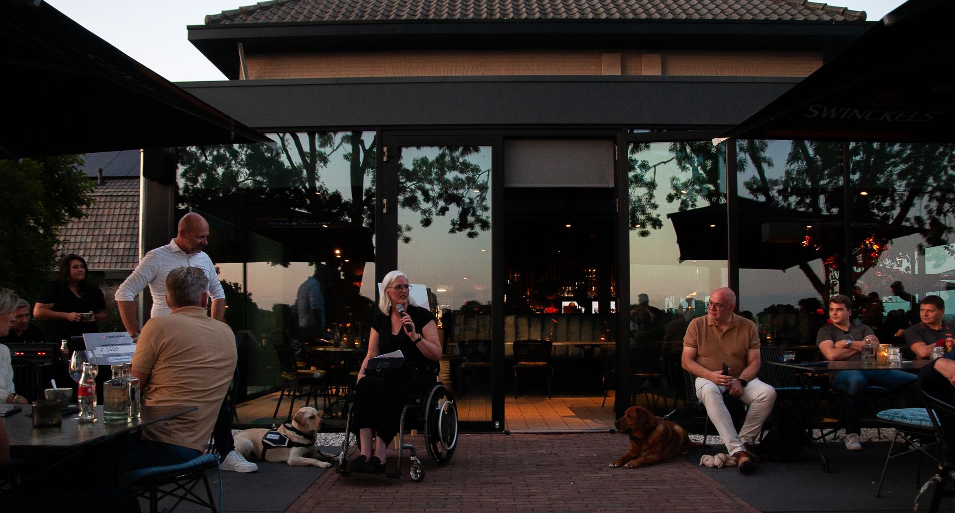 Mensen verzamelden zich bij schemering op een terras, sommigen zaten, anderen stonden bij een gebouw met glazen ramen die de zonsondergang weerspiegelden.