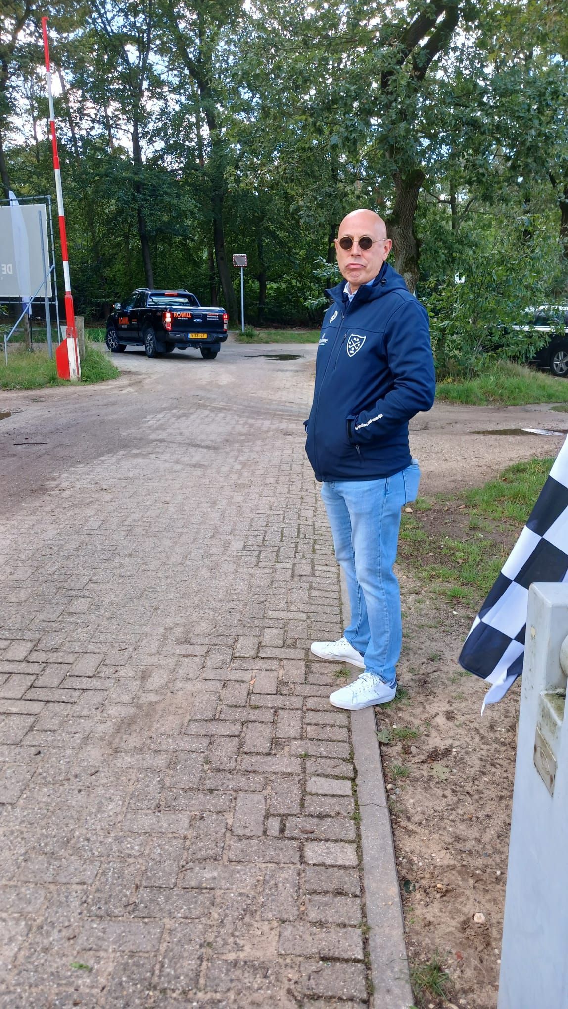 Een man in een blauwe jas en spijkerbroek staat naast een weg, met een spoorwegovergang op de achtergrond.