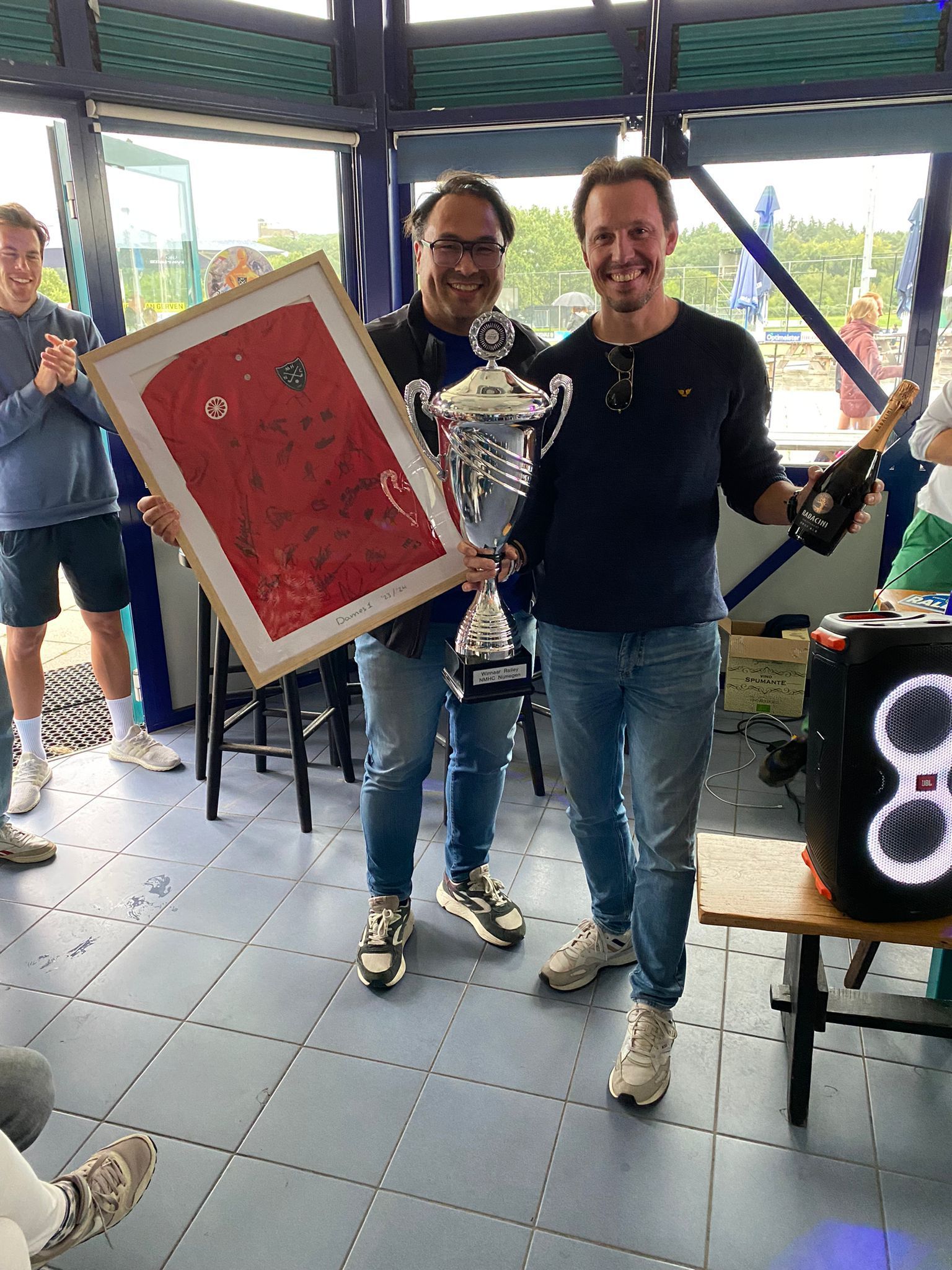 Twee mannen houden een ingelijst shirt en een trofee vast en vieren feest. Een van hen ontkurkt een champagnefles. Locatie: buitenfeest.