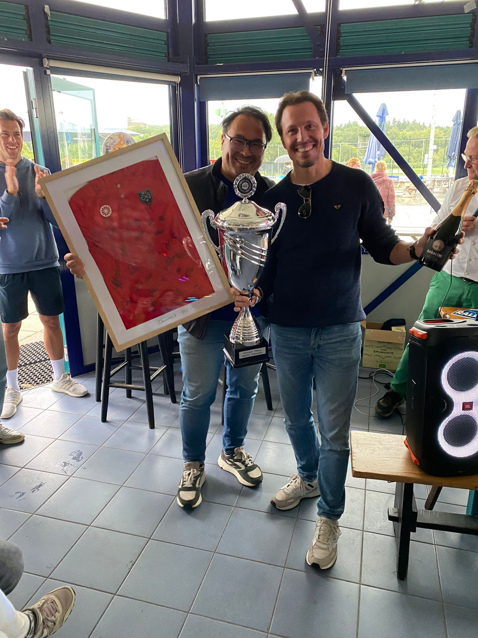 Twee mannen vieren hun overwinning en houden een trofee en een ingelijst kunstwerk vast. Een van hen heeft een fles champagne in zijn hand.