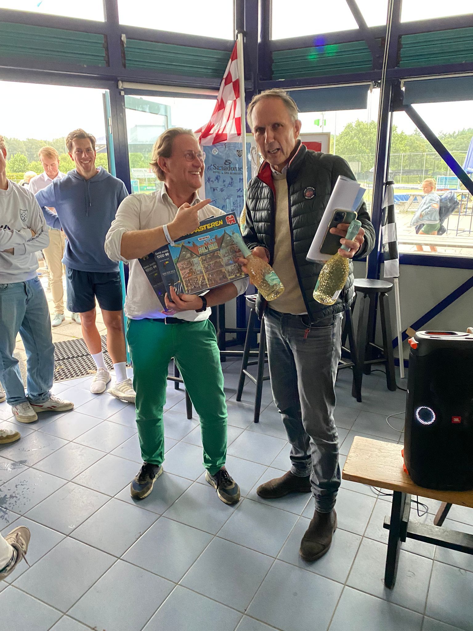 Twee mensen vieren feest binnenshuis; de een heeft een cadeau vast, de ander een fles champagne, en ze glimlachen.