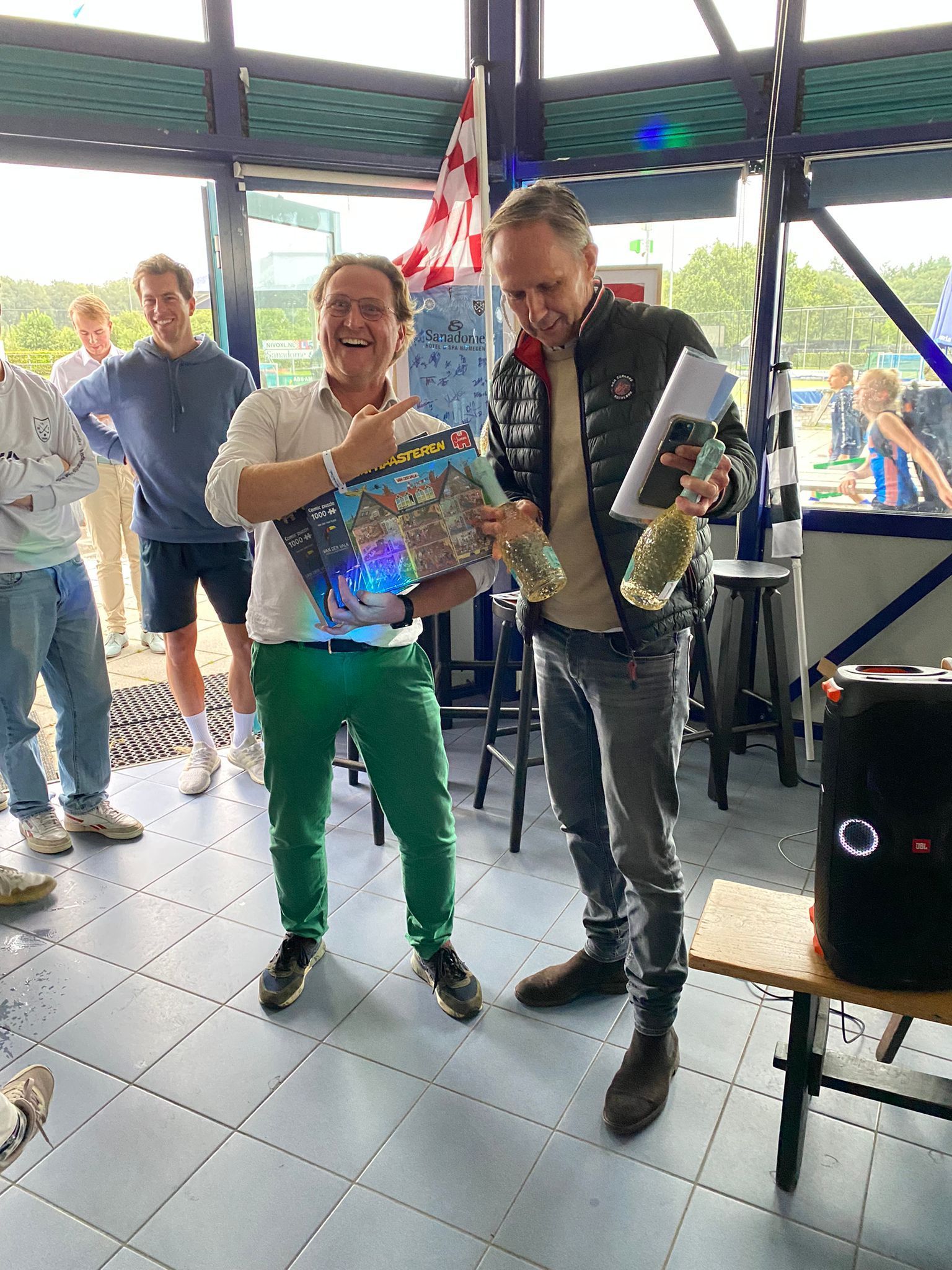Twee mannen vieren feest binnenshuis. De ene houdt een ingelijste foto vast, de andere champagne en een certificaat.