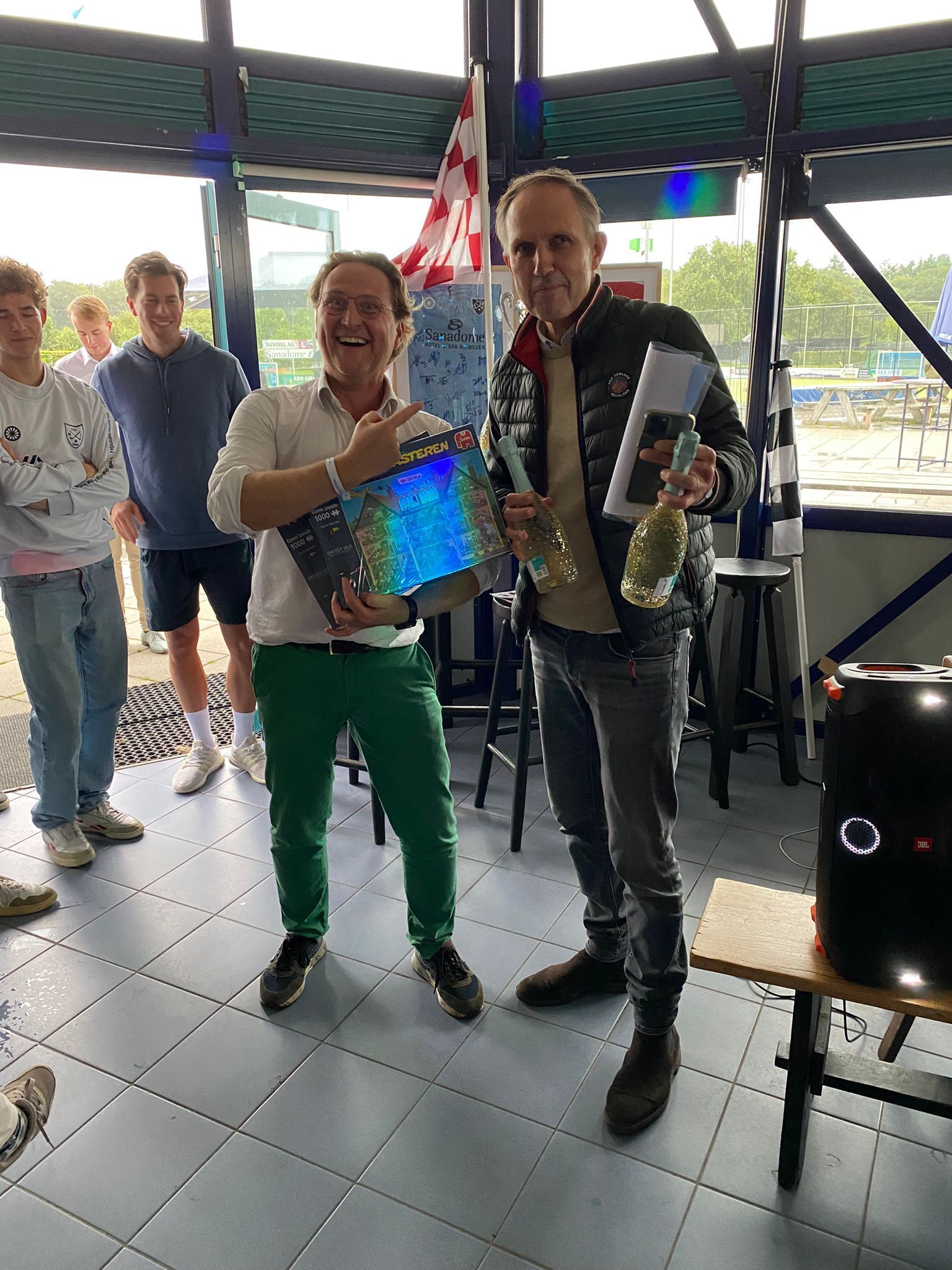 Binnen vieren twee mannen feest met een trofee en een fles champagne; op de achtergrond zijn andere mensen te zien.