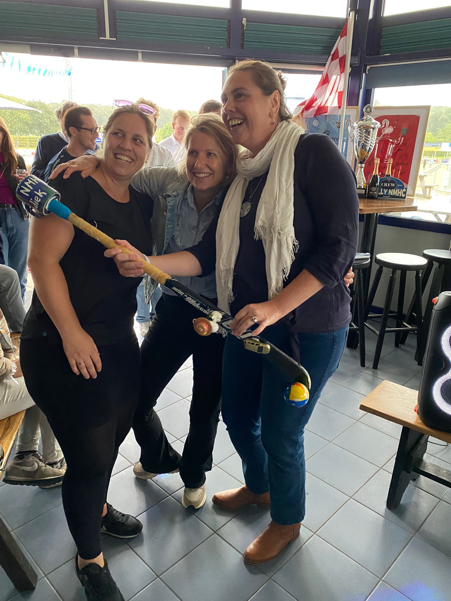 Drie vrouwen, lachend en met een hockeystick en een bal in hun handen, vieren feest binnenshuis.