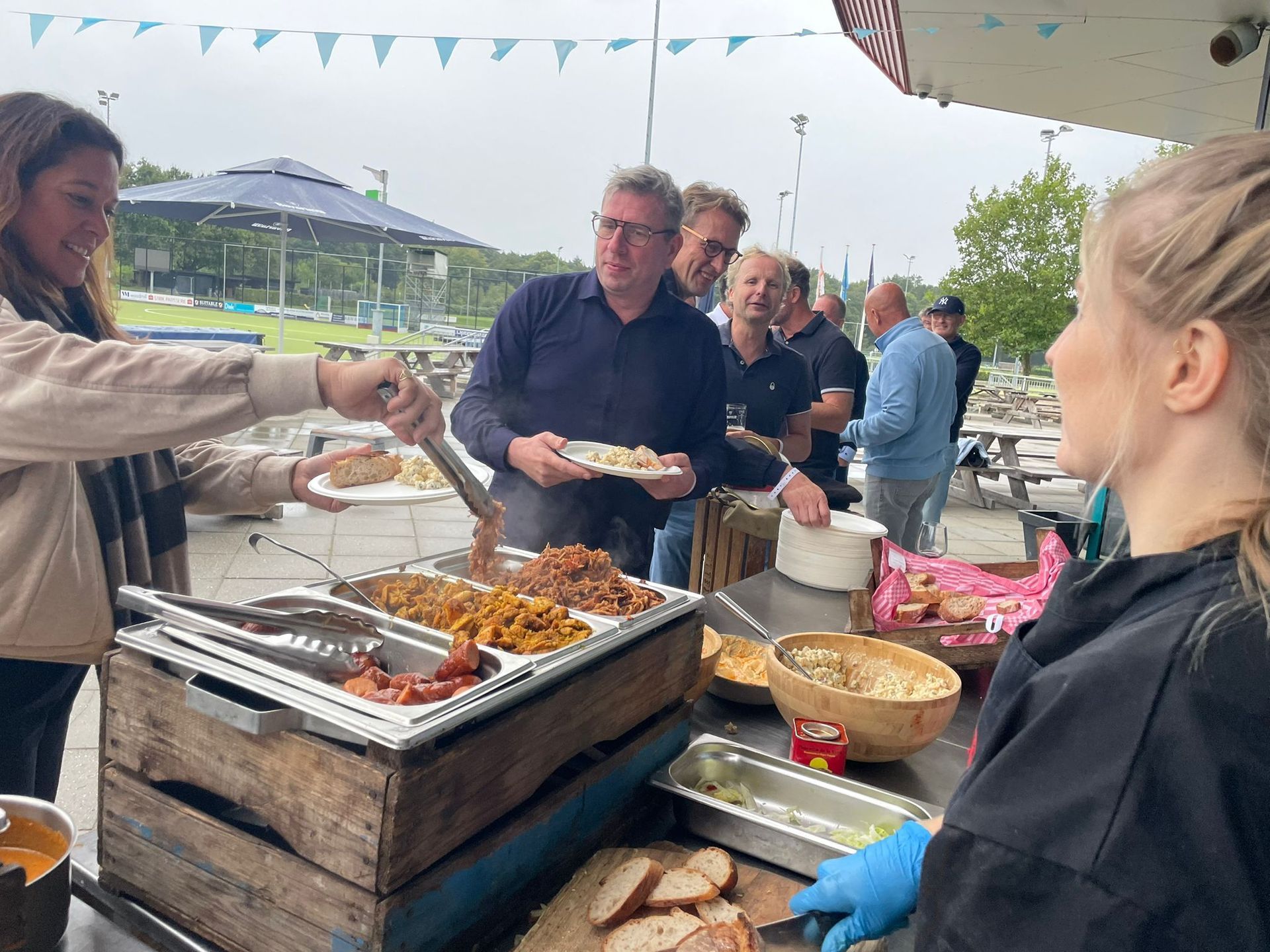 Mensen die eten serveren en geserveerd krijgen tijdens een evenement in de buitenlucht. De ober gebruikt een tang; gasten staan ​​in de rij met hun borden.