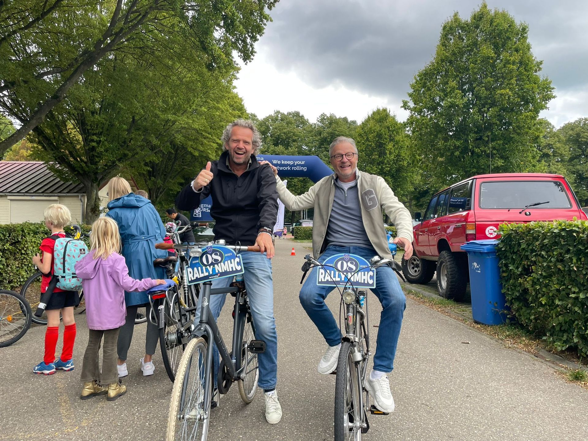 Twee mannen op fietsen steken hun duim omhoog; anderen staan ​​erbij. Buiten, vlakbij bomen, een blauwe boog.
