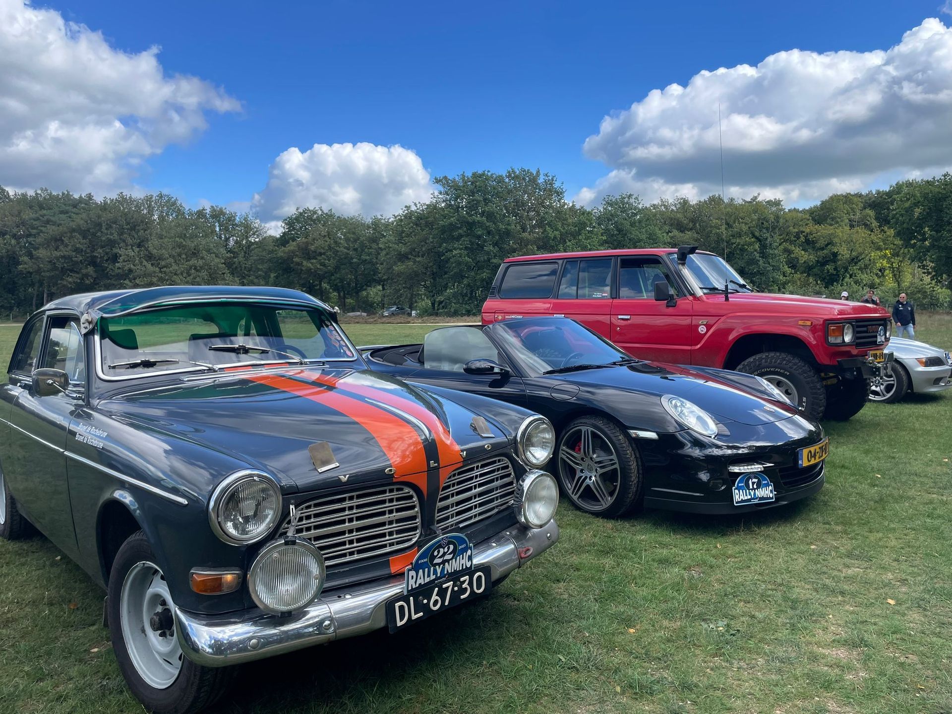 Klassieke auto's geparkeerd op het gras onder een blauwe hemel: een groene Volvo, een zwarte Porsche en een rode terreinwagen.