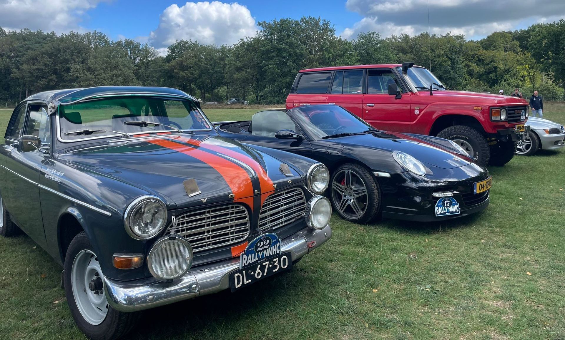 Klassieke auto's geparkeerd op het gras: een Volvo, een Porsche en een rode SUV onder een zonnige hemel.