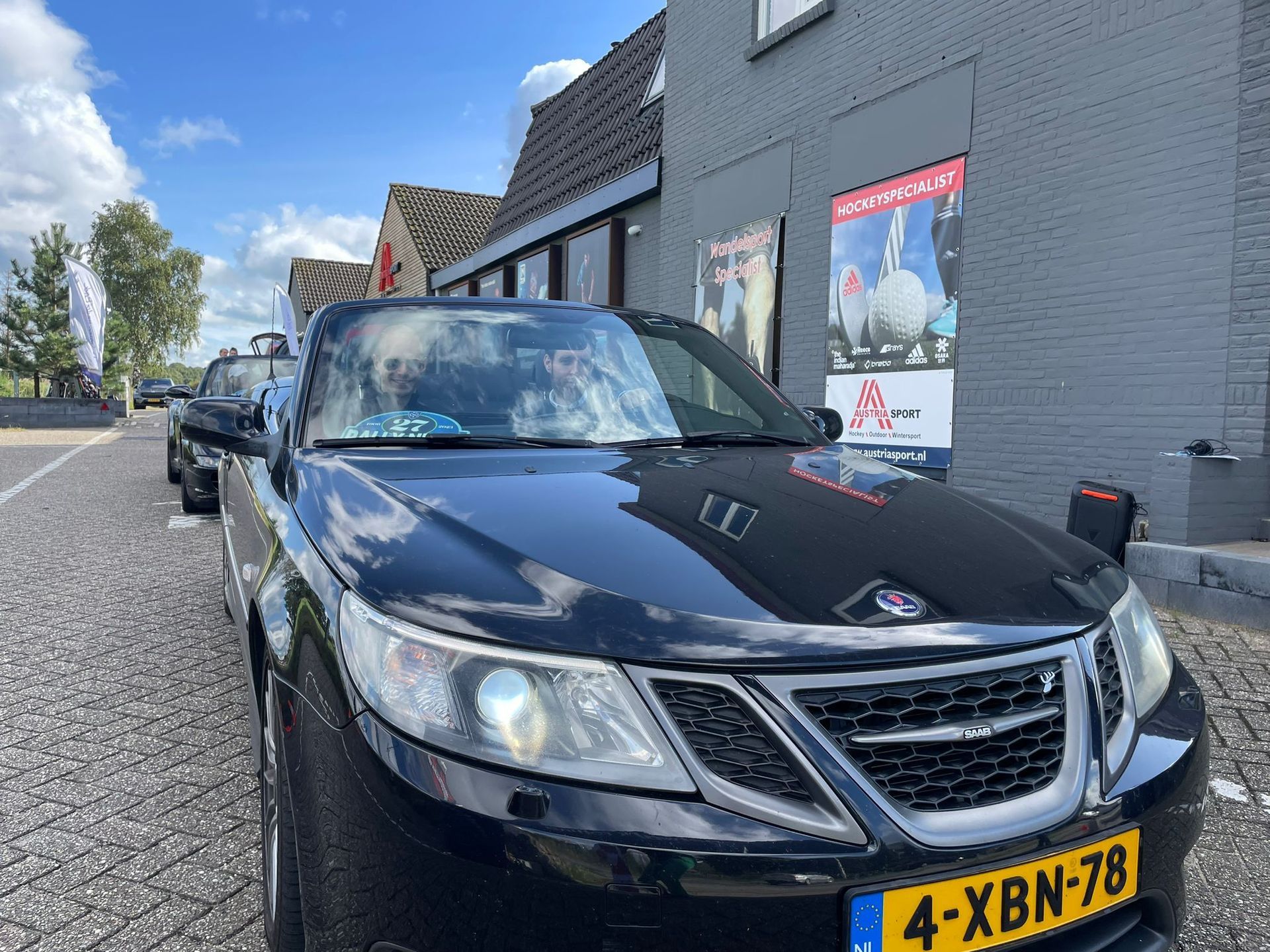 Een zwarte Saab staat geparkeerd op een straat met kinderkopjes voor een grijs gebouw, onder een gedeeltelijk bewolkte hemel.