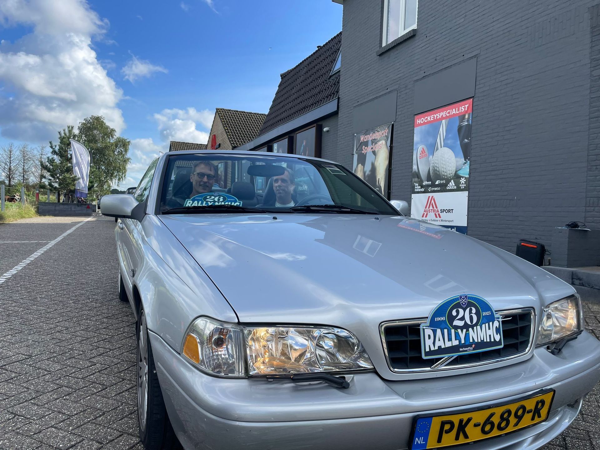 Een zilverkleurige Volvo met twee inzittenden staat geparkeerd voor een grijs gebouw. ​​Op de grille is een rally-embleem te zien.