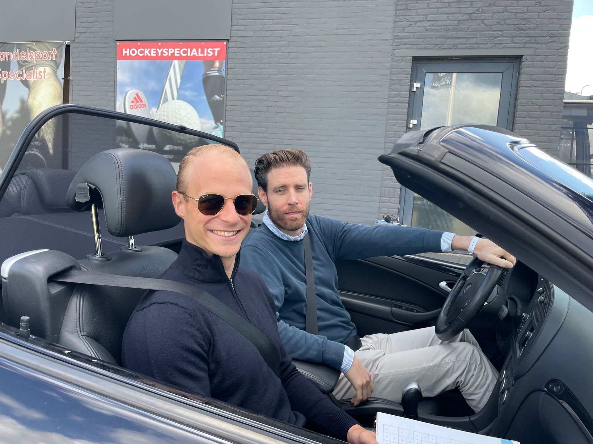Twee mannen in een cabriolet, lachend. De ene draagt ​​een zonnebril, de andere bestuurt. Buiten, overdag.