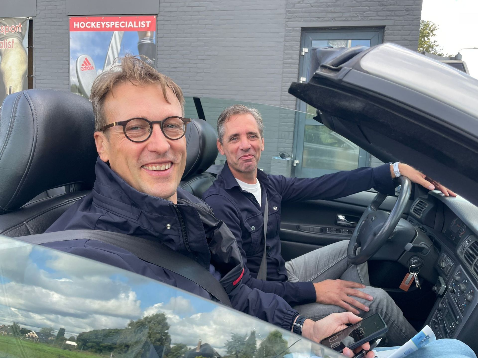 Twee mannen in een cabriolet, één achter het stuur. Beiden glimlachen, buiten. Grijze auto, overdag.
