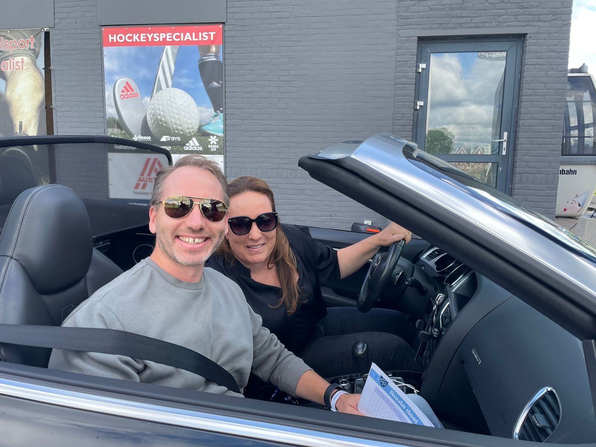 Twee mensen in een cabriolet, lachend. Buiten, vlakbij een gebouw met reclame voor sportartikelen.