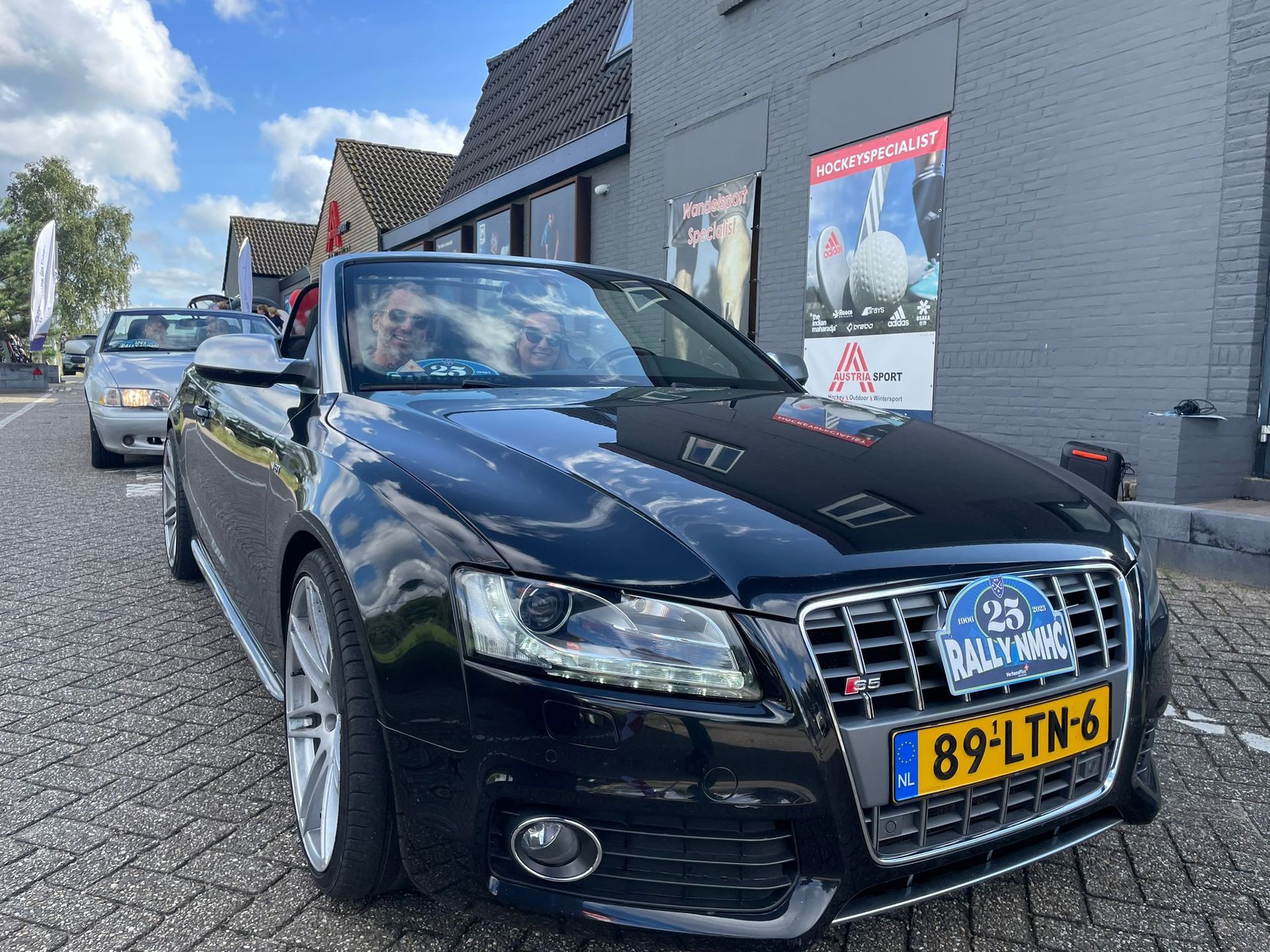 Een zwarte Audi cabriolet met kenteken 89-LTM-6 staat voor een gebouw met mensen erin.