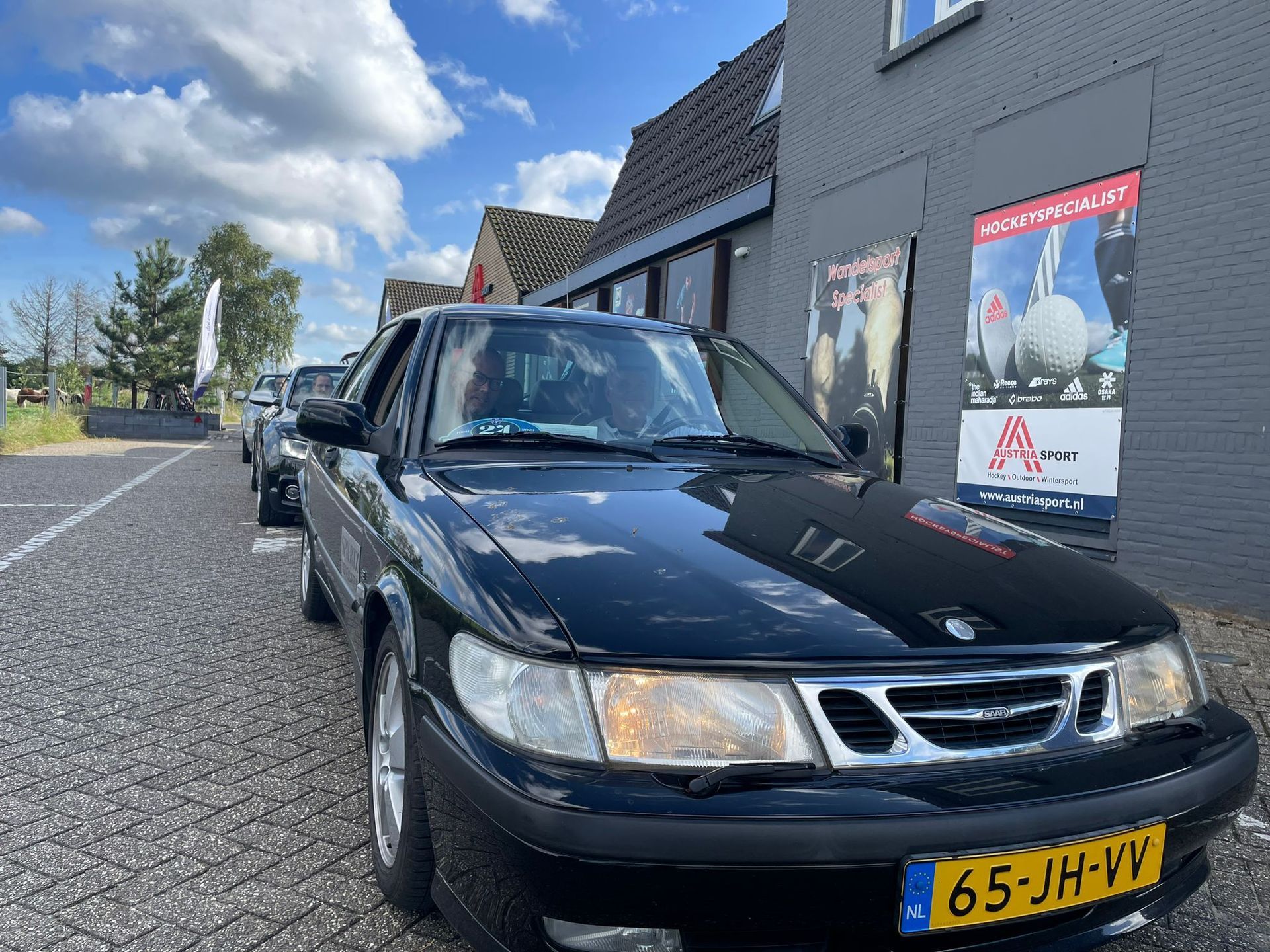 Een zwarte Saab 9-3 staat geparkeerd voor een gebouw met posters, onder een bewolkte hemel.