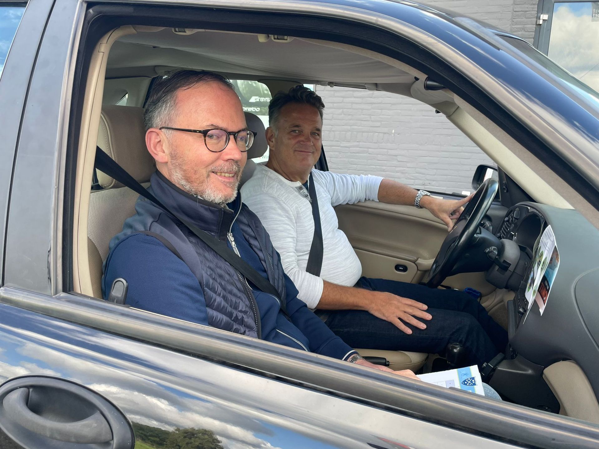 Twee mannen in een auto. De ene rijdt, de andere lacht. Overdag, natuurlijk licht.