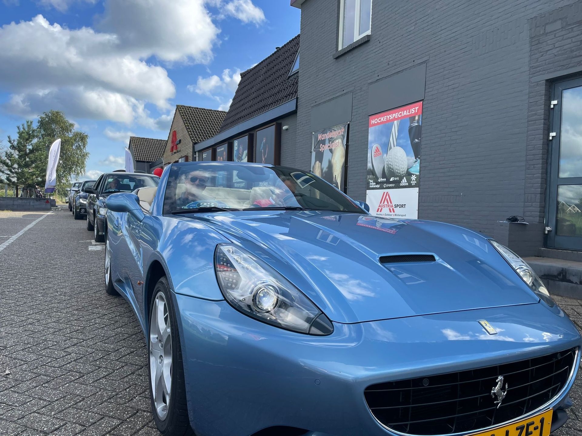 Een blauwe Ferrari cabriolet staat geparkeerd voor een gebouw, tussen andere auto's.