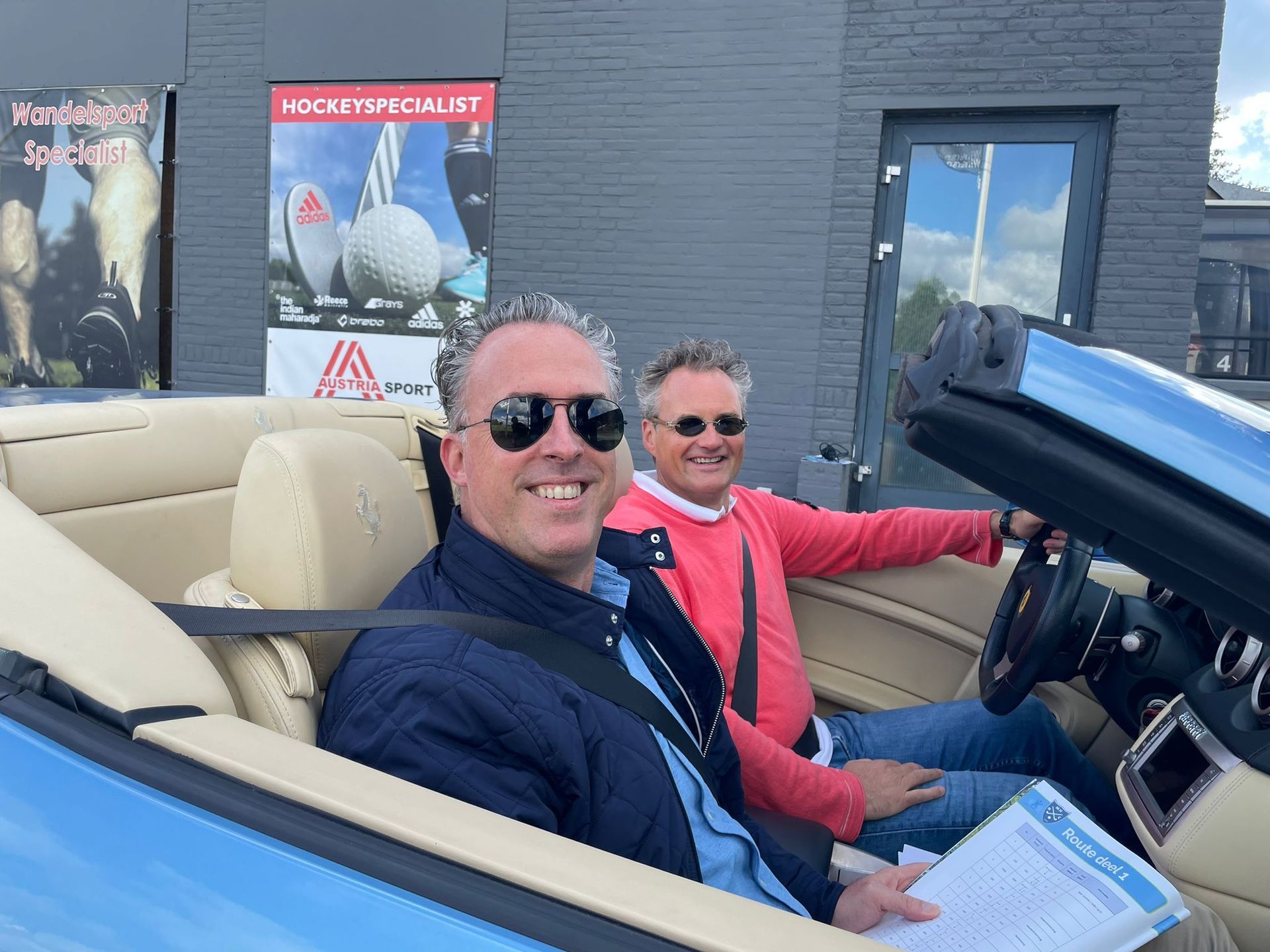 Twee mannen glimlachen vanuit een lichtblauwe cabriolet. De ene draagt ​​een zonnebril en een blauwe jas, de andere een rode trui.