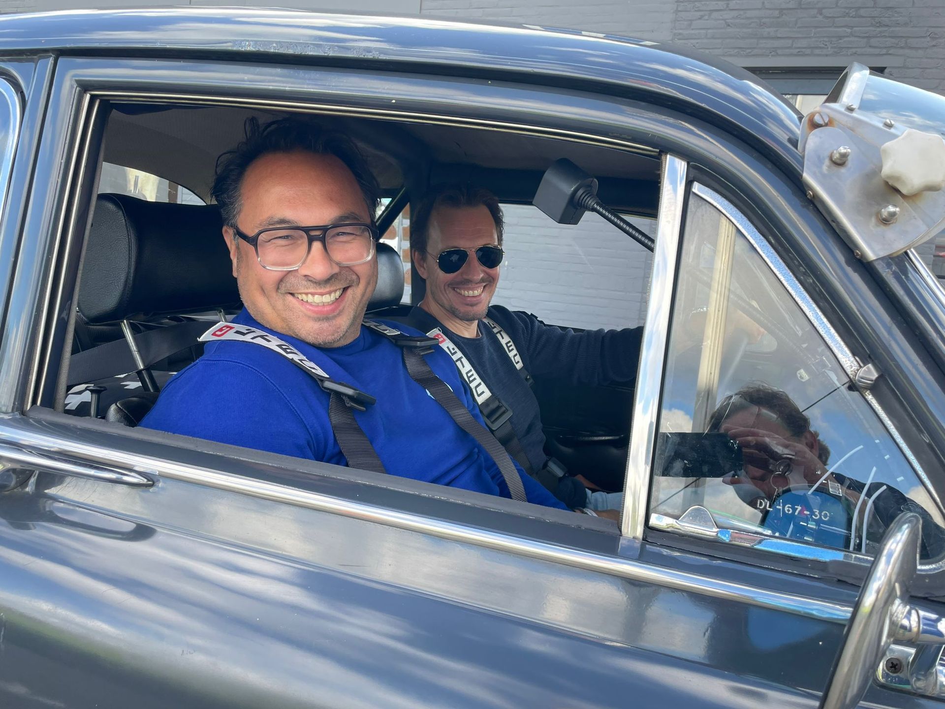 Twee mensen in een oldtimer, lachend. De ene draagt ​​een bril en een blauw shirt, de andere een zwart shirt.
