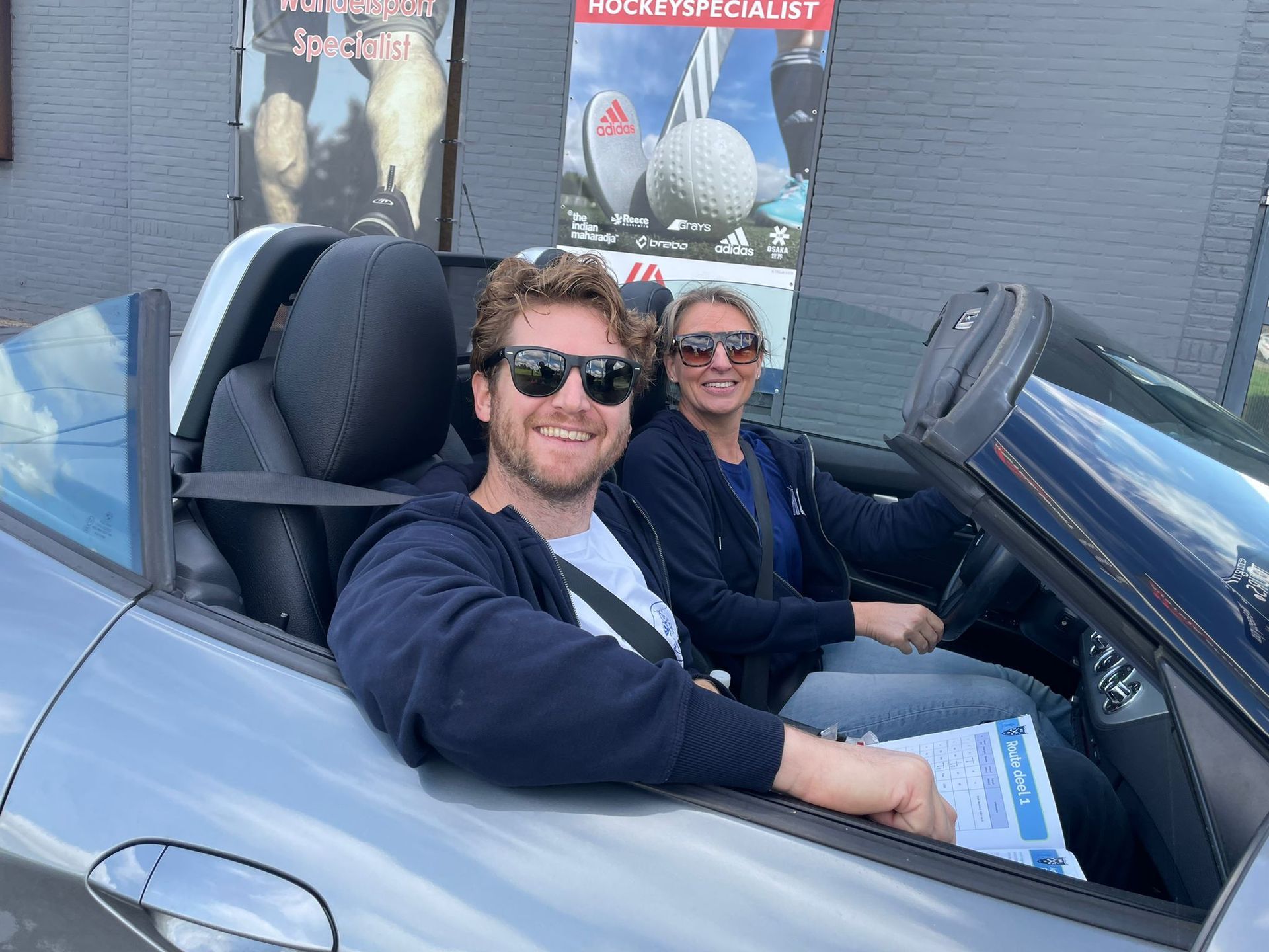 Twee mensen met zonnebrillen zitten in een zilverkleurige cabriolet. Ze glimlachen, rijden en houden papieren vast.