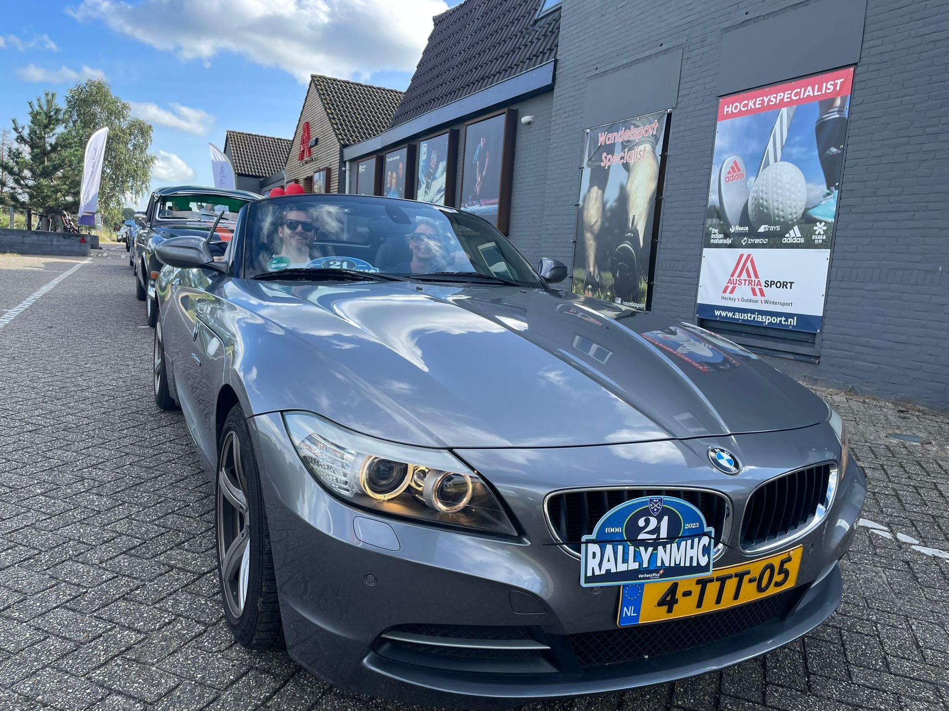Een grijze BMW Z4 cabriolet met een rallykentekenplaat staat geparkeerd voor een gebouw met posters.