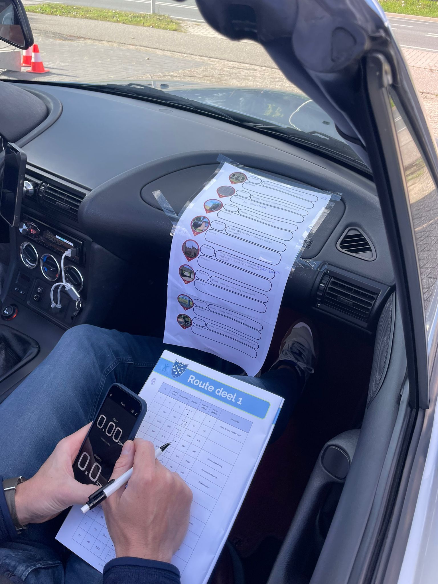 Persoon in auto die een stopwatch gebruikt, aantekeningen maakt op een klembord en een ander vel papier met afbeeldingen heeft liggen.