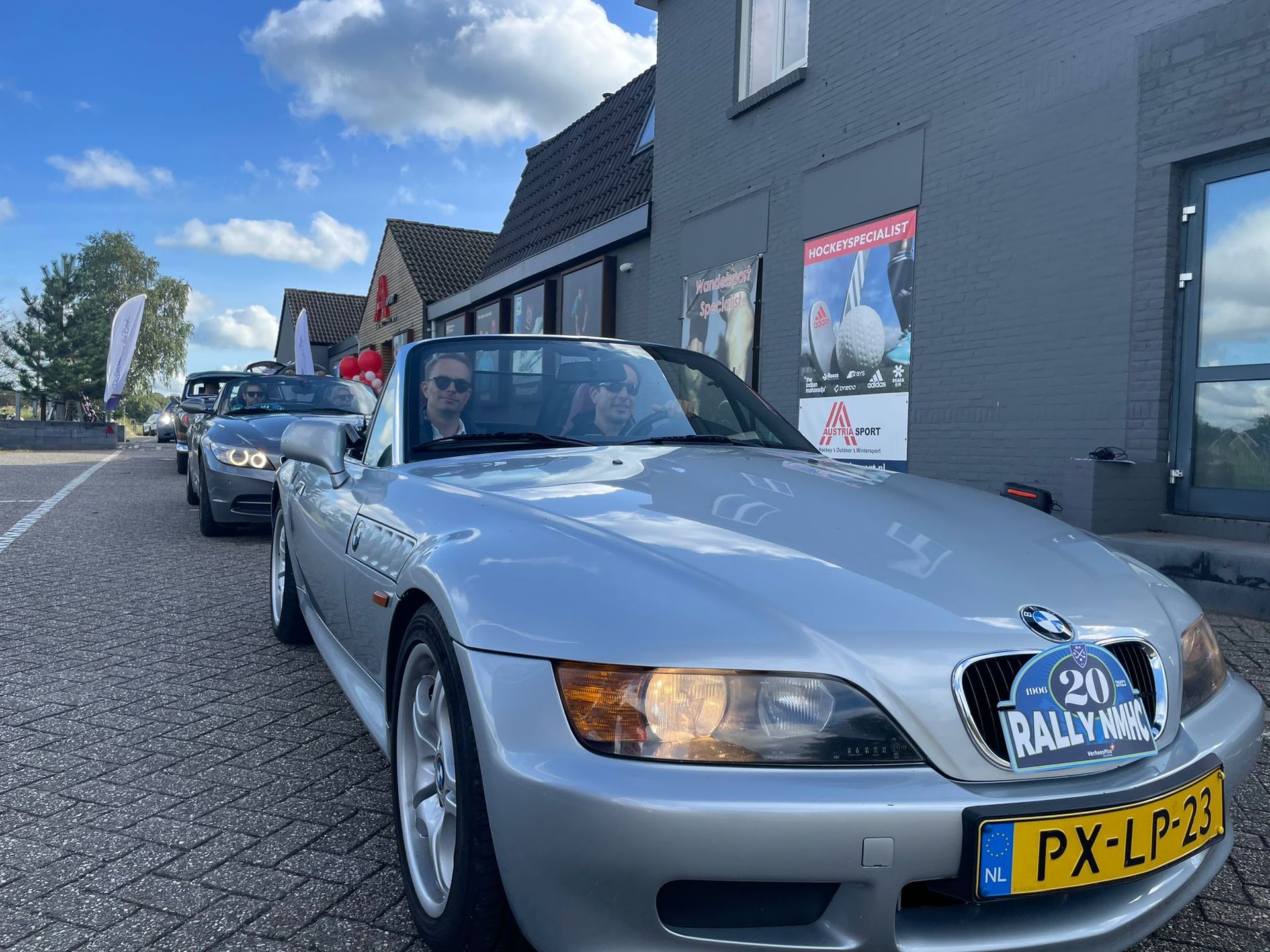 Een zilverkleurige BMW Z3 cabriolet staat in een rij met andere auto's geparkeerd bij een gebouw. ​​Met een rallysticker op de voorkant.