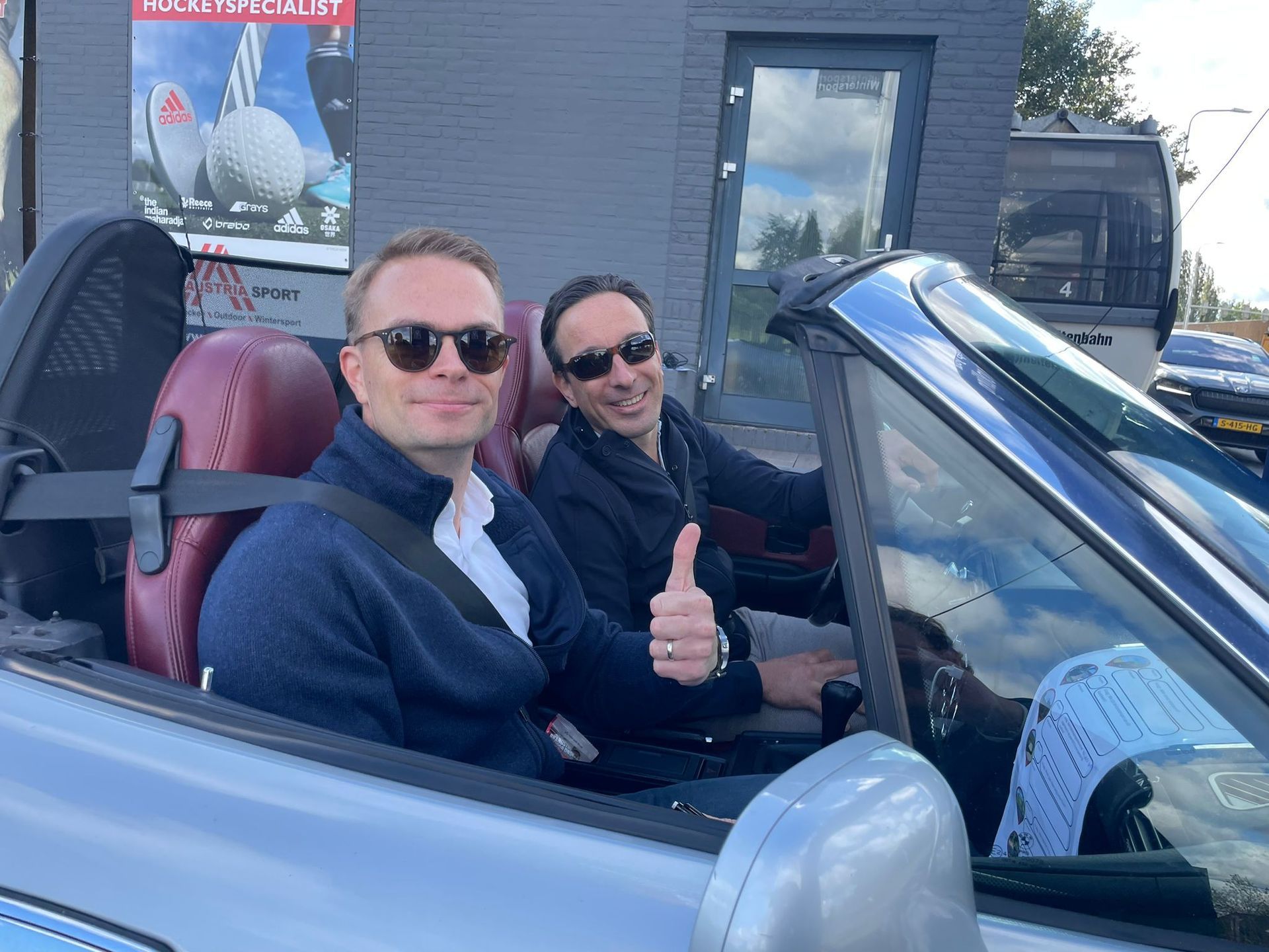 Twee mannen in een zilverkleurige cabriolet. Een van hen steekt zijn duim omhoog; beiden dragen een zonnebril. Buitenopname.
