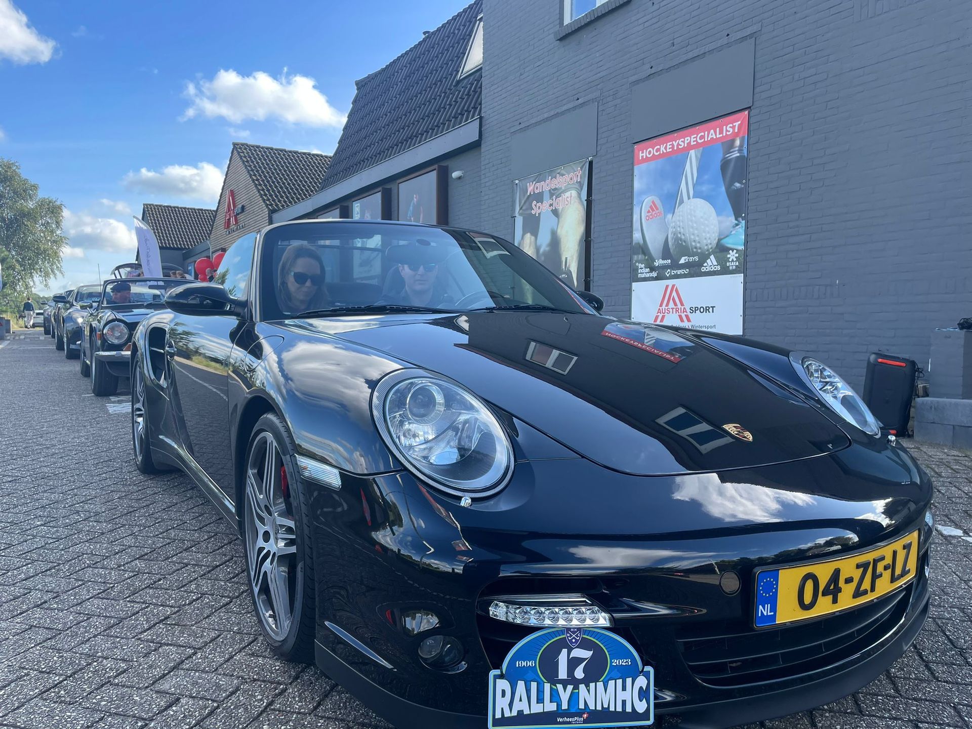 Zwarte Porsche cabriolet met passagiers, geparkeerd tussen andere auto's; exterieur, overdag.