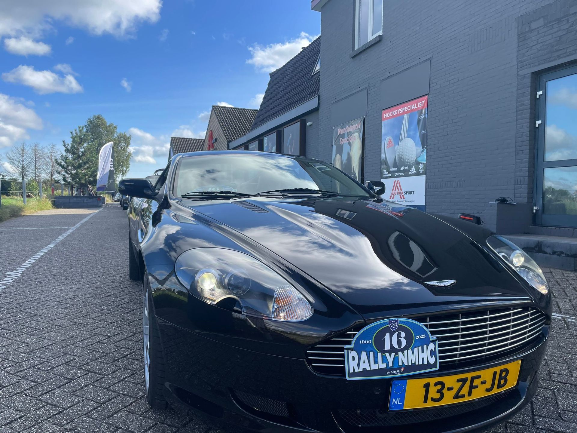 Een zwarte Aston Martin sportwagen staat geparkeerd voor een grijs gebouw. ​​Het kenteken luidt 