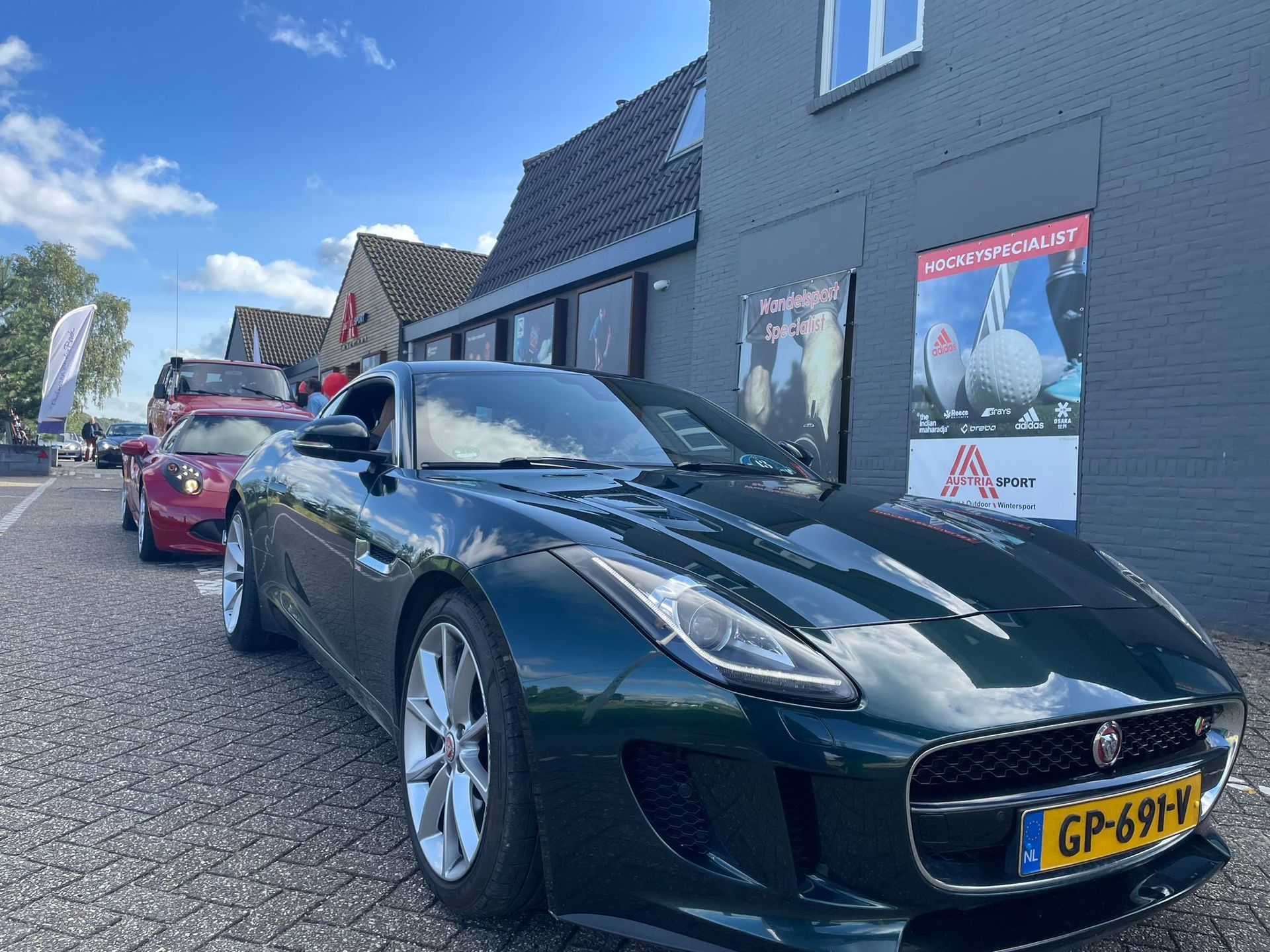 Een groene Jaguar sportwagen staat geparkeerd voor een gebouw met reclameborden die met golf te maken hebben; op de achtergrond zijn andere auto's te zien.