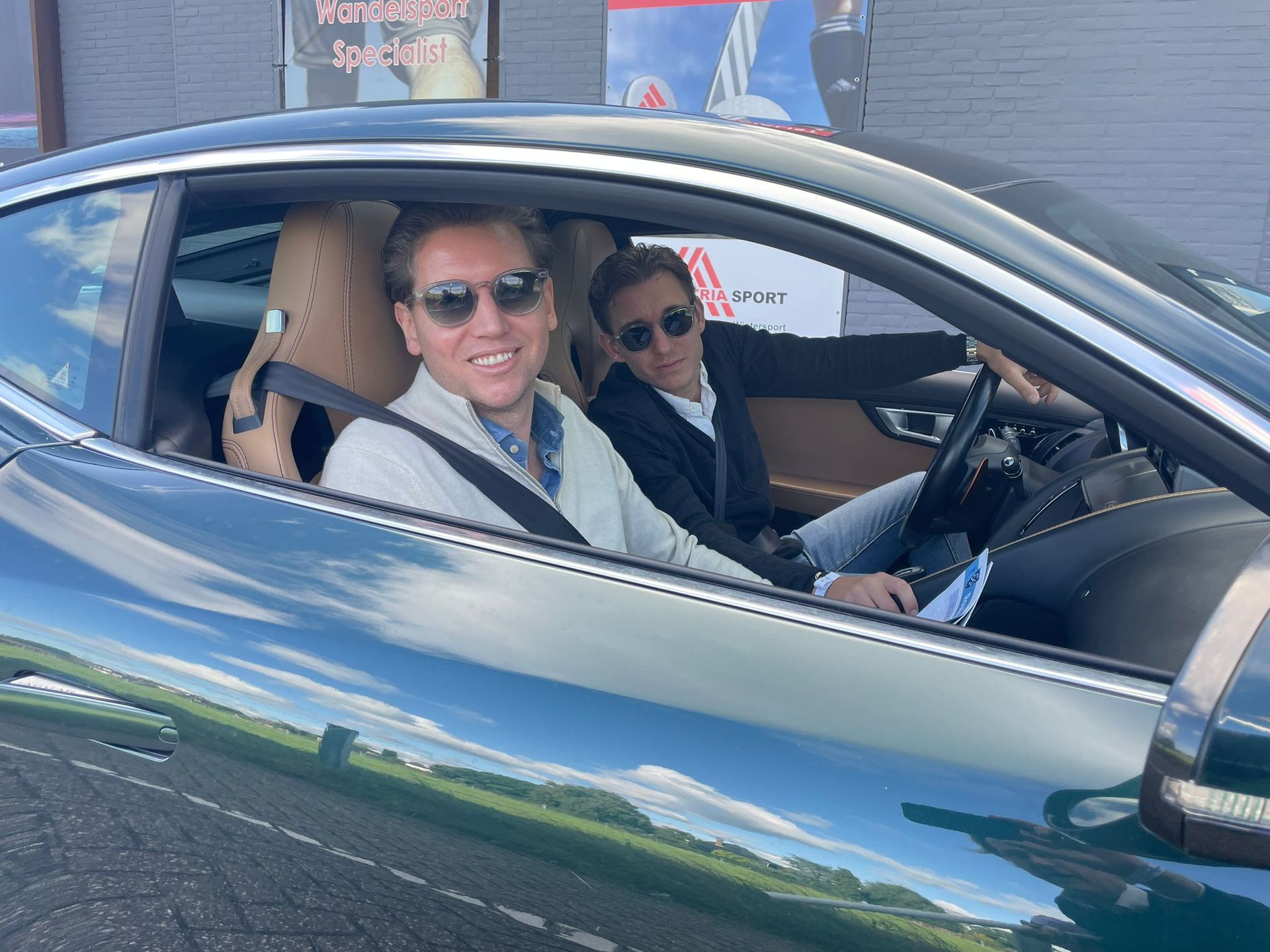 Twee mannen in een groene sportwagen. De ene bestuurt de auto, de andere lacht. Zonnige dag.