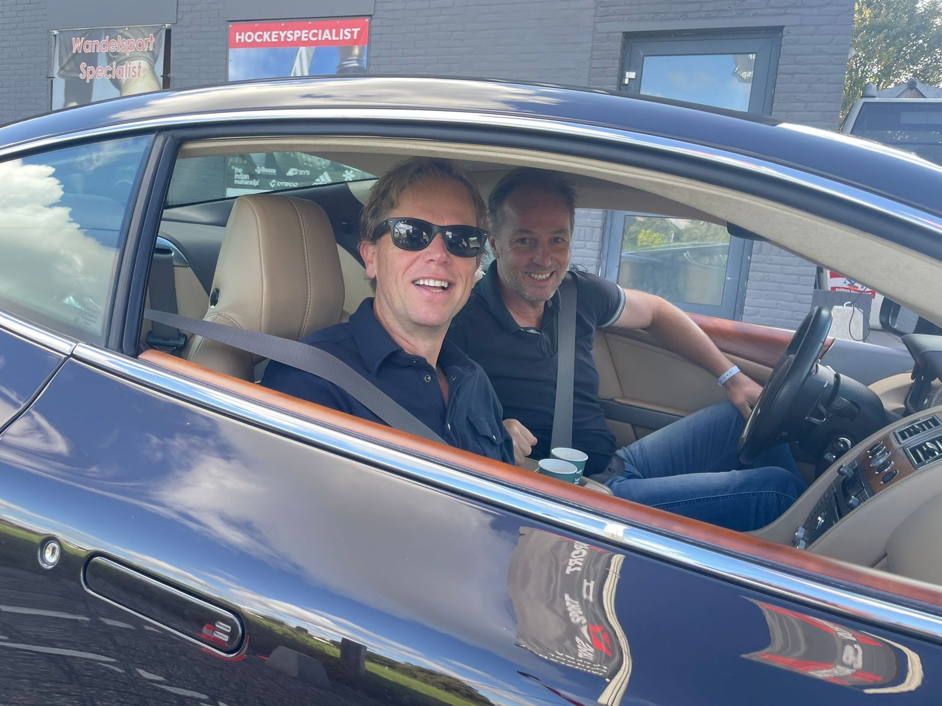 Twee mannen glimlachen in een donkerblauwe sportwagen met beige interieur.