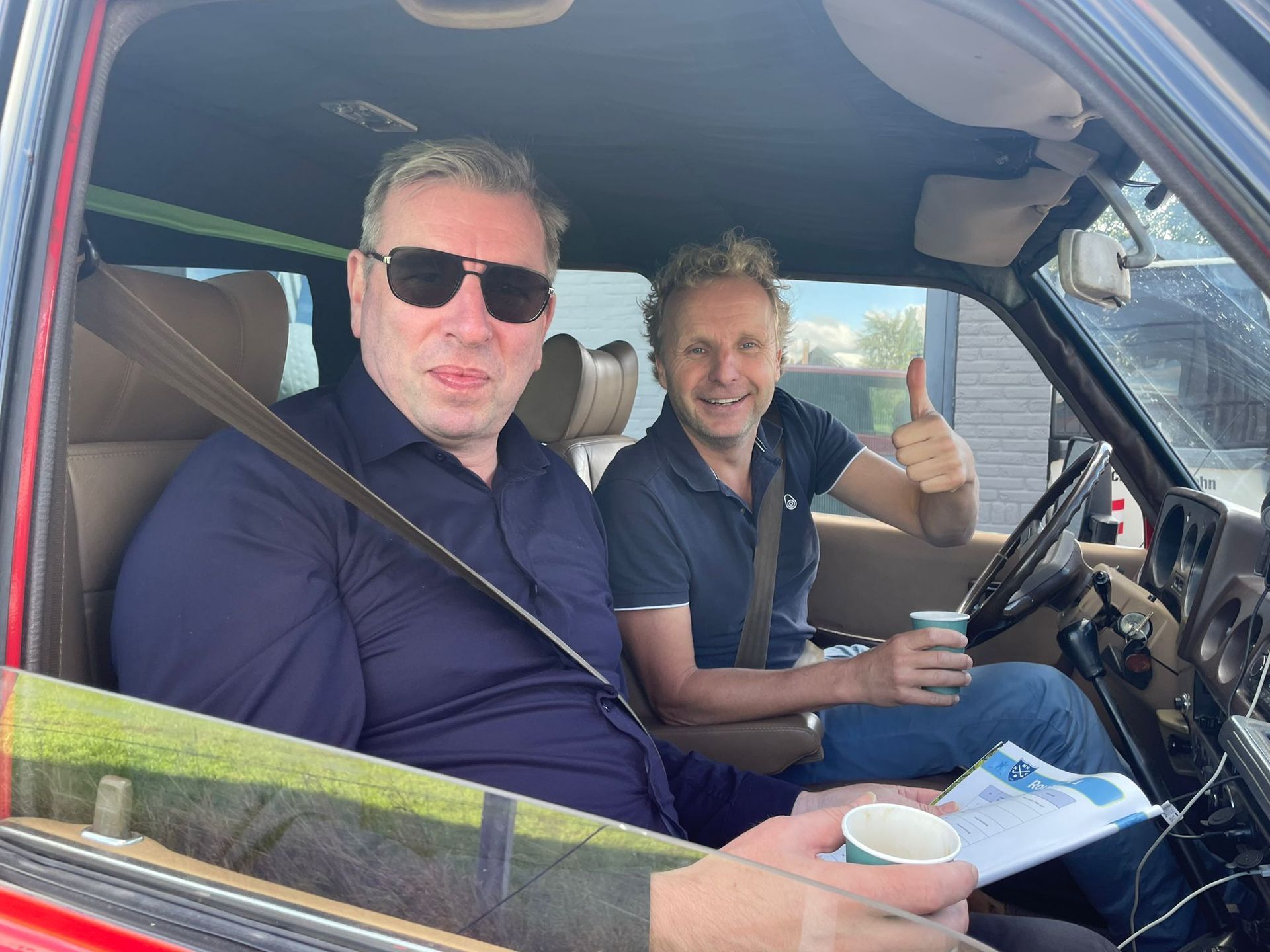 Twee mannen in een rode auto. De ene draagt ​​een zonnebril, de andere steekt zijn duim omhoog terwijl hij een koffiebeker vasthoudt.