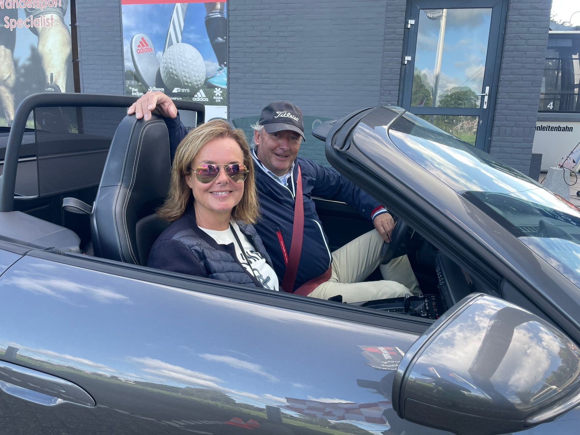 Een stel in een grijze cabriolet lacht naar de camera.