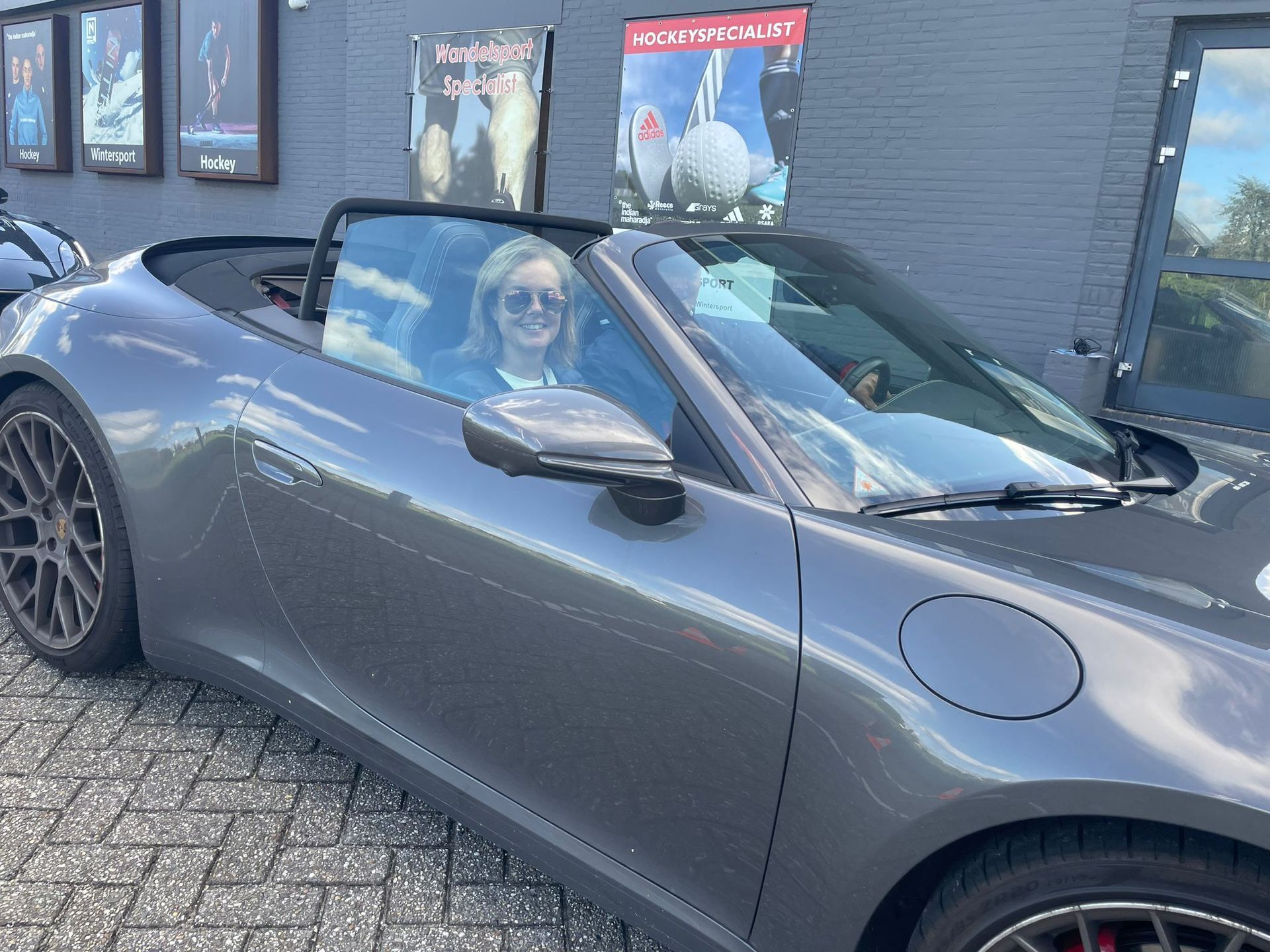 Een vrouw glimlacht vanuit een grijze cabriolet die voor een gebouw geparkeerd staat.