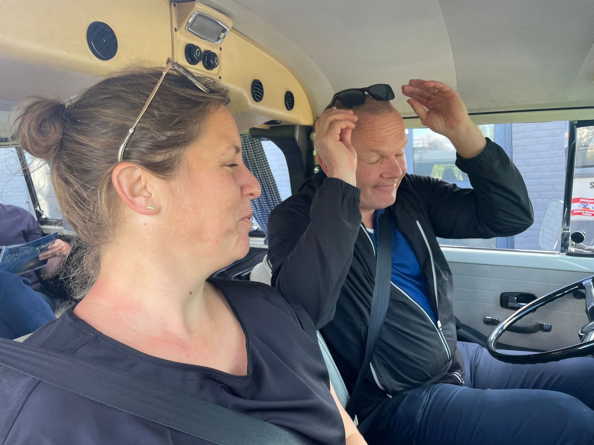 Een vrouw en een man in een lichtgekleurde auto. De man zet zijn zonnebril recht, de vrouw kijkt weg.