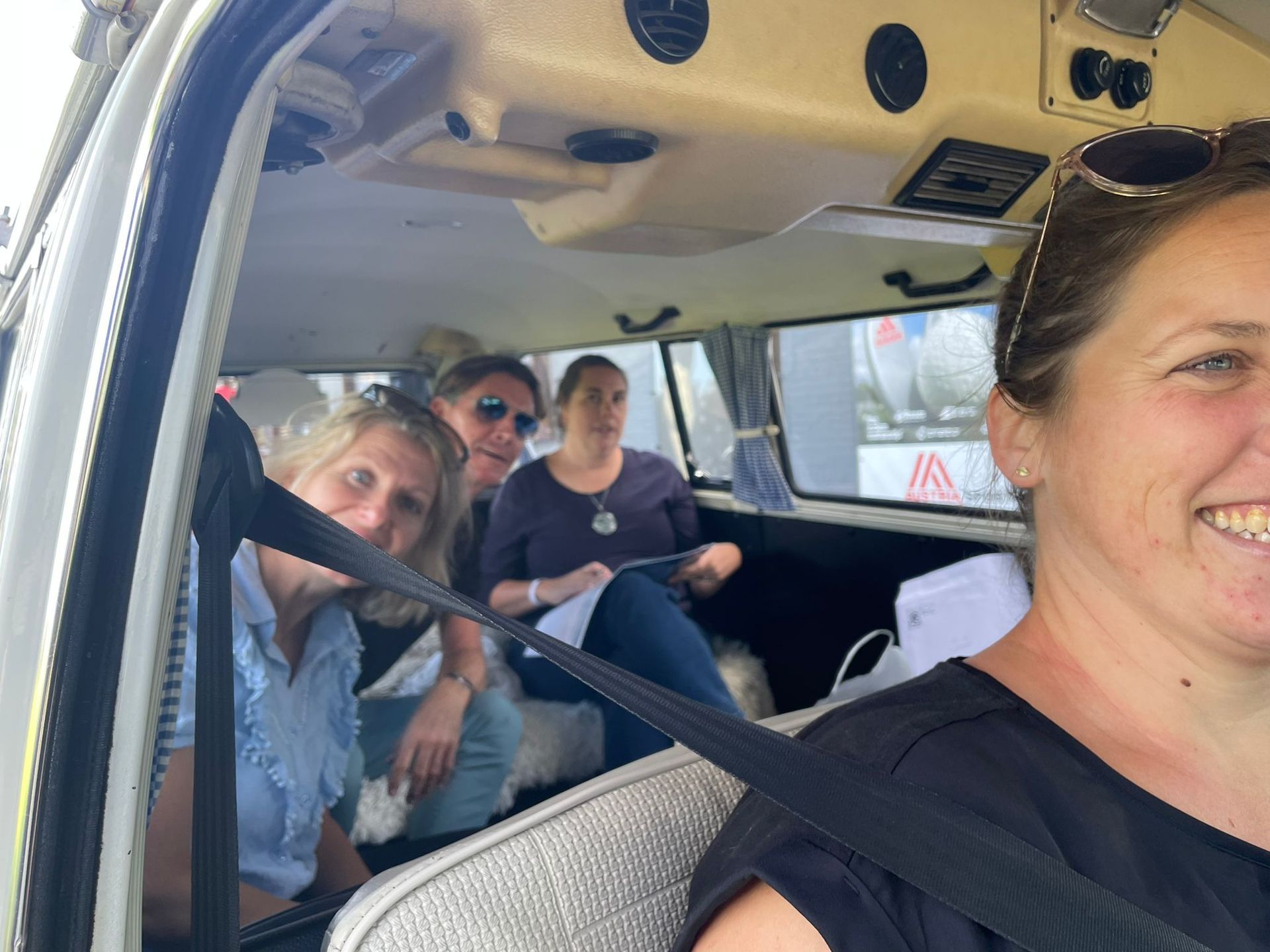 Vier vrouwen glimlachen in een vintage camper. De bestuurster draagt ​​een zonnebril, de anderen zitten met papieren in hun handen.