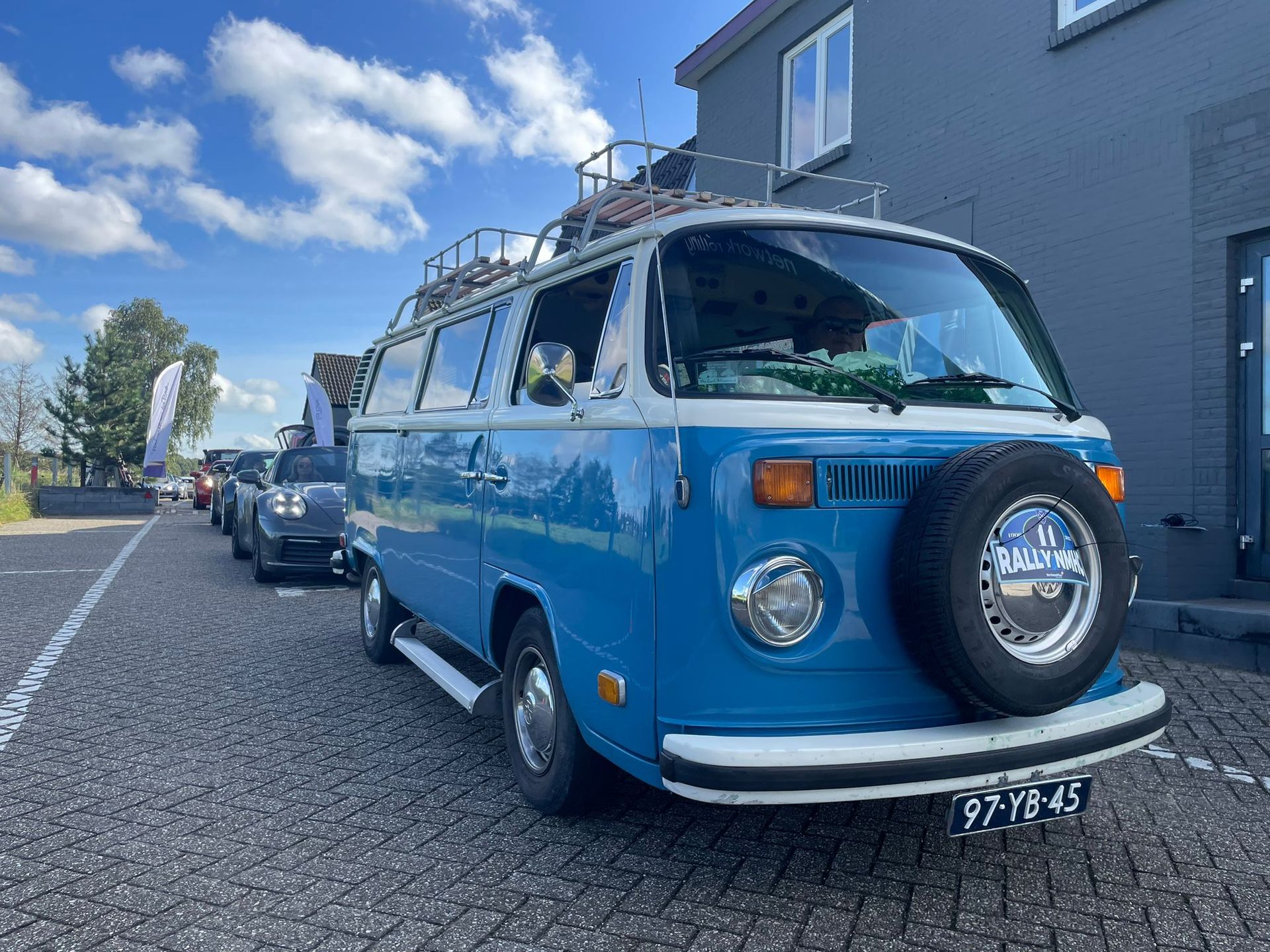 Blauw-witte Volkswagenbus met imperiaal geparkeerd op een kasseienweg. Reservewiel vooraan gemonteerd.