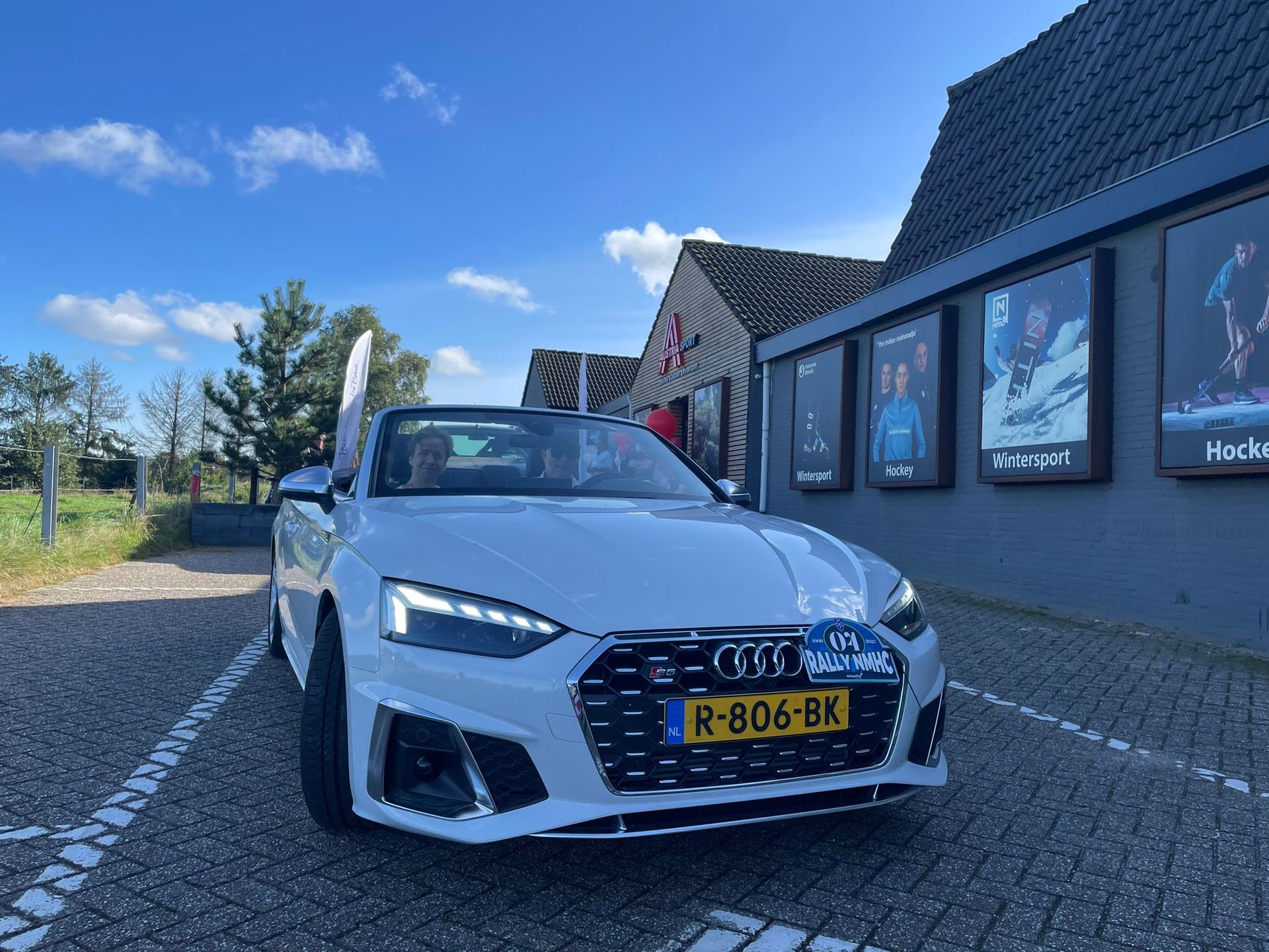 Een witte Audi cabriolet staat geparkeerd voor een gebouw met filmposters op een zonnige dag.