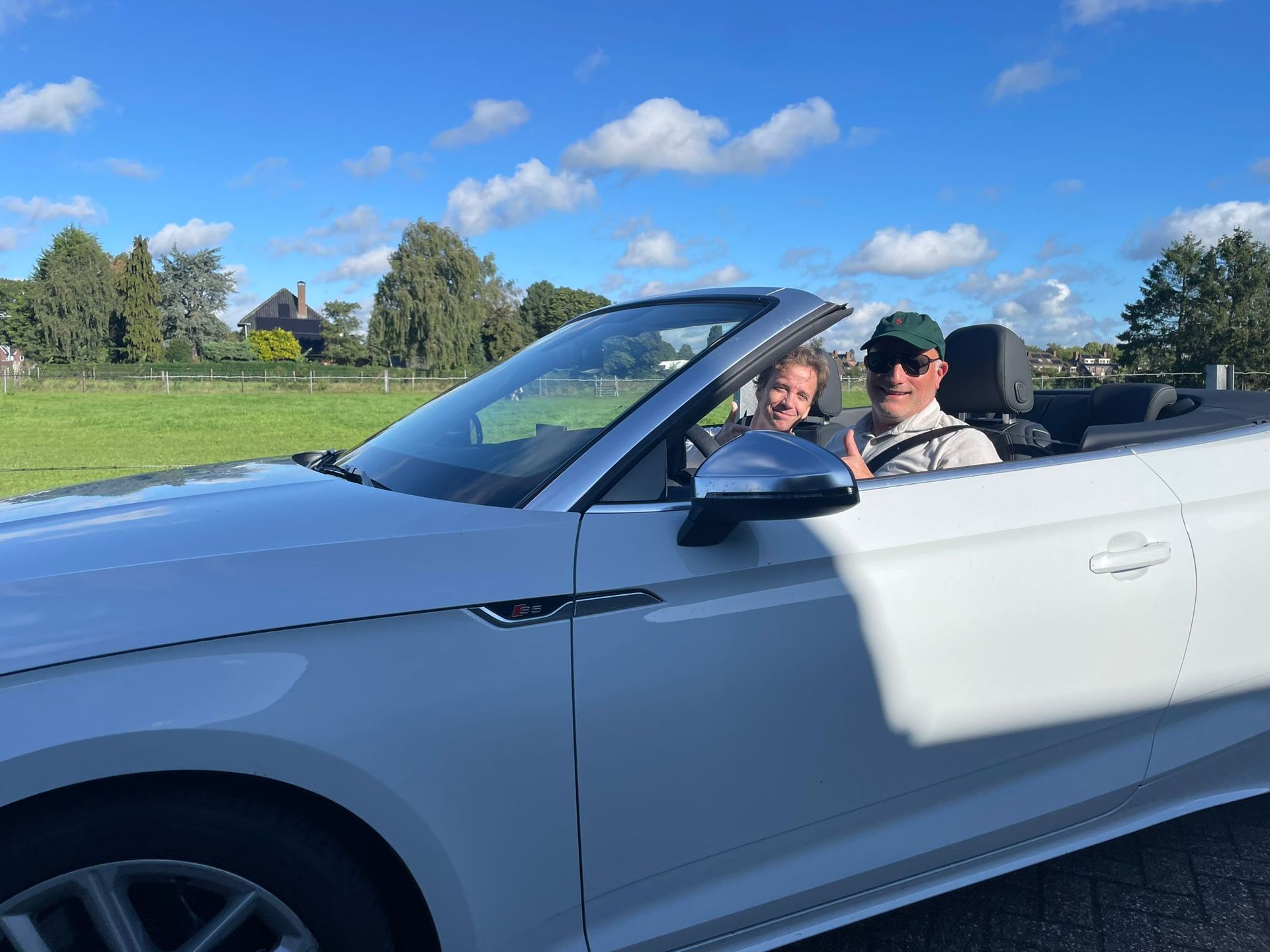Een witte cabriolet met twee inzittenden. Een persoon met een groene pet steekt zijn duim omhoog, met een jong persoon op de passagiersstoel.