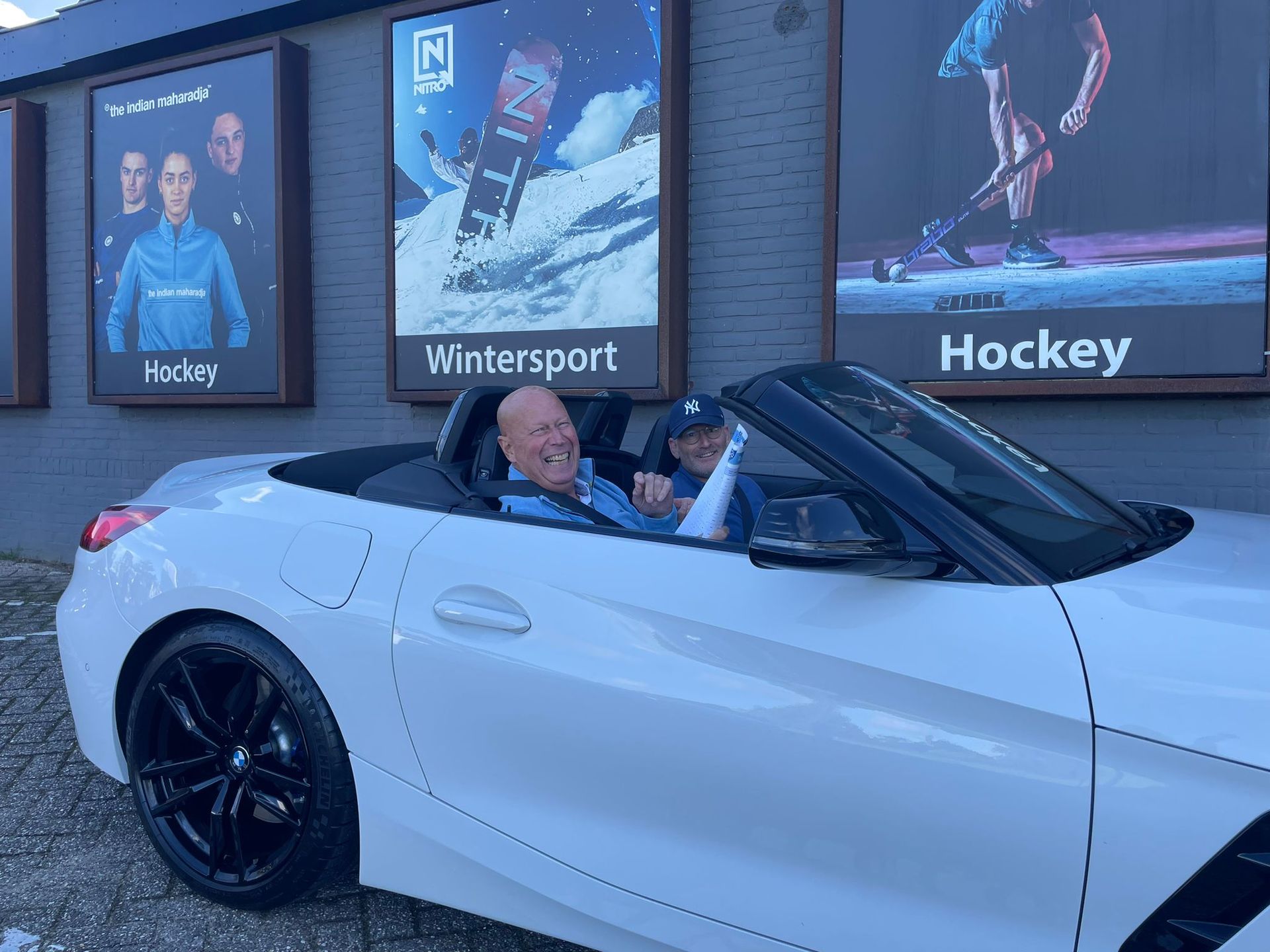 Man en kind in een witte cabriolet. Op de achtergrond zijn borden met 'Hockey', 'Wintersport' en 'Hockey' te zien.