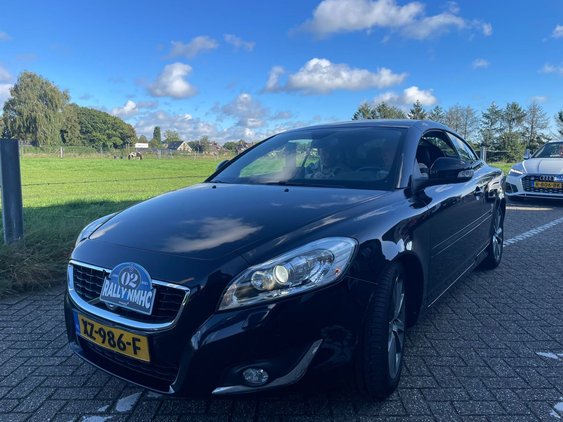Een zwarte Volvo coupé staat geparkeerd op een stoep, met op de achtergrond een grasveld en bomen.