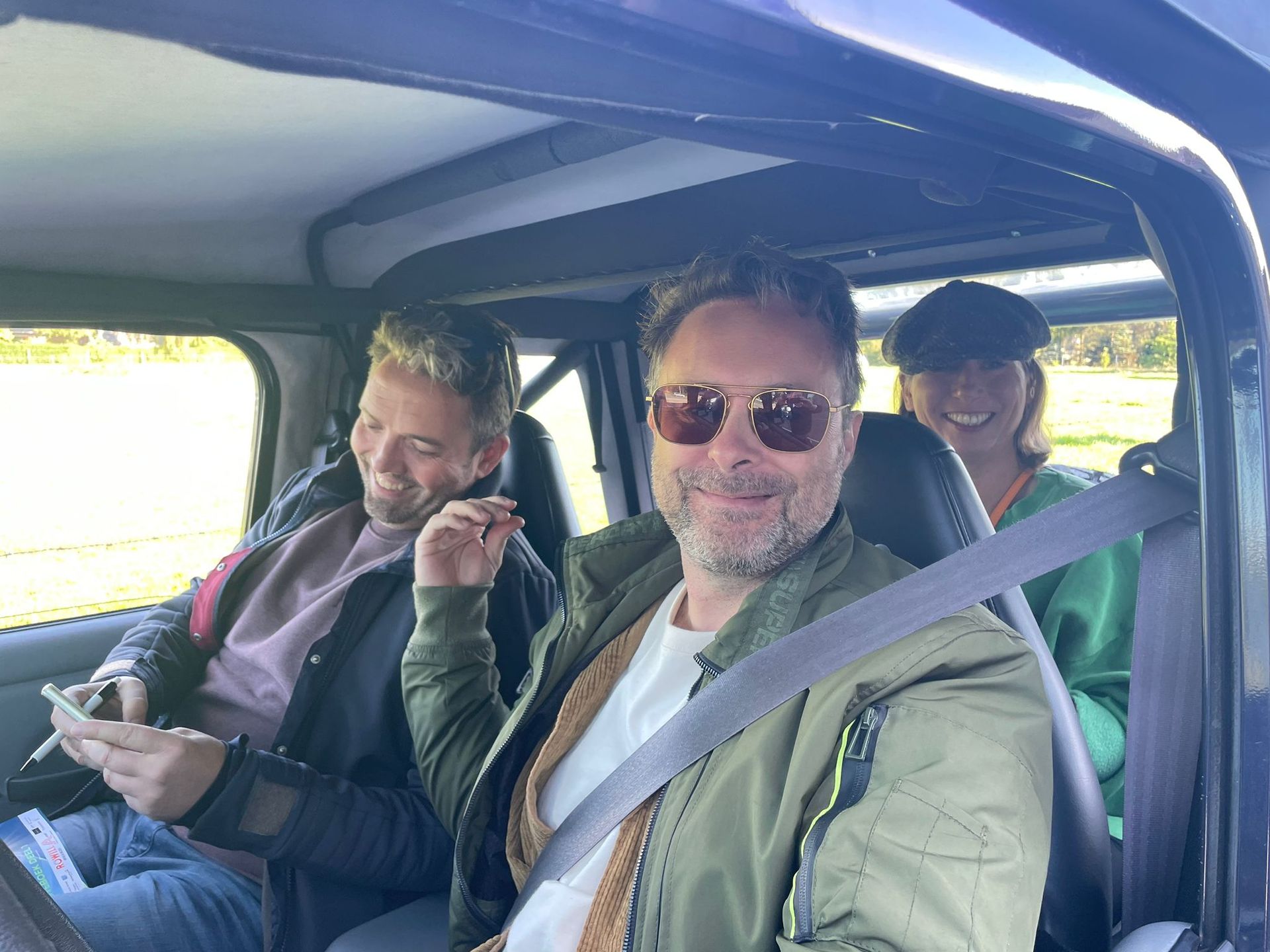 Drie mensen in een Jeep: een man met een zonnebril lacht, een ander kijkt op zijn telefoon en een derde met een pet lacht ook.