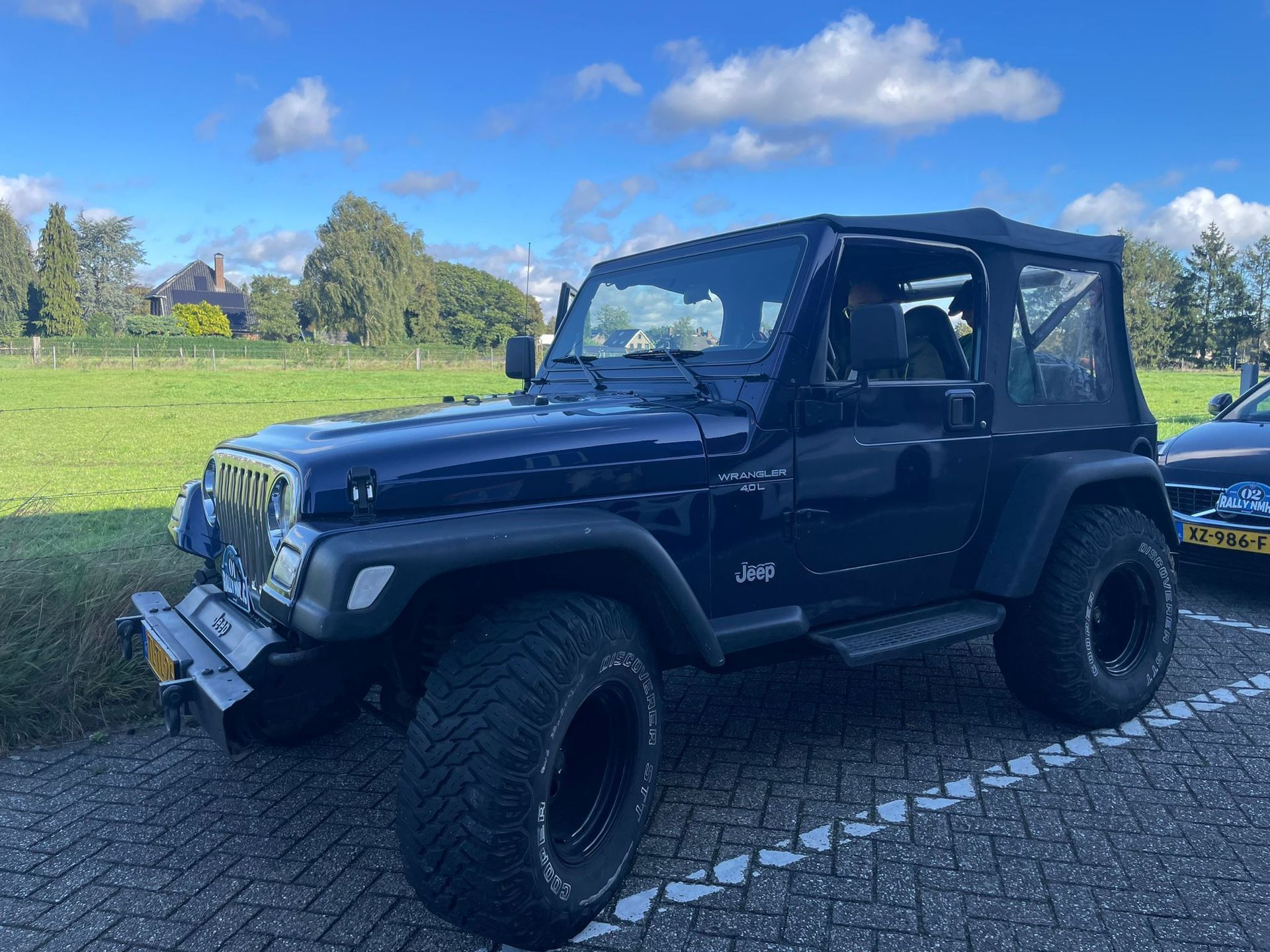 Een donkerblauwe Jeep Wrangler met softtop staat geparkeerd op asfalt; op de achtergrond zijn een grasveld en bomen te zien.
