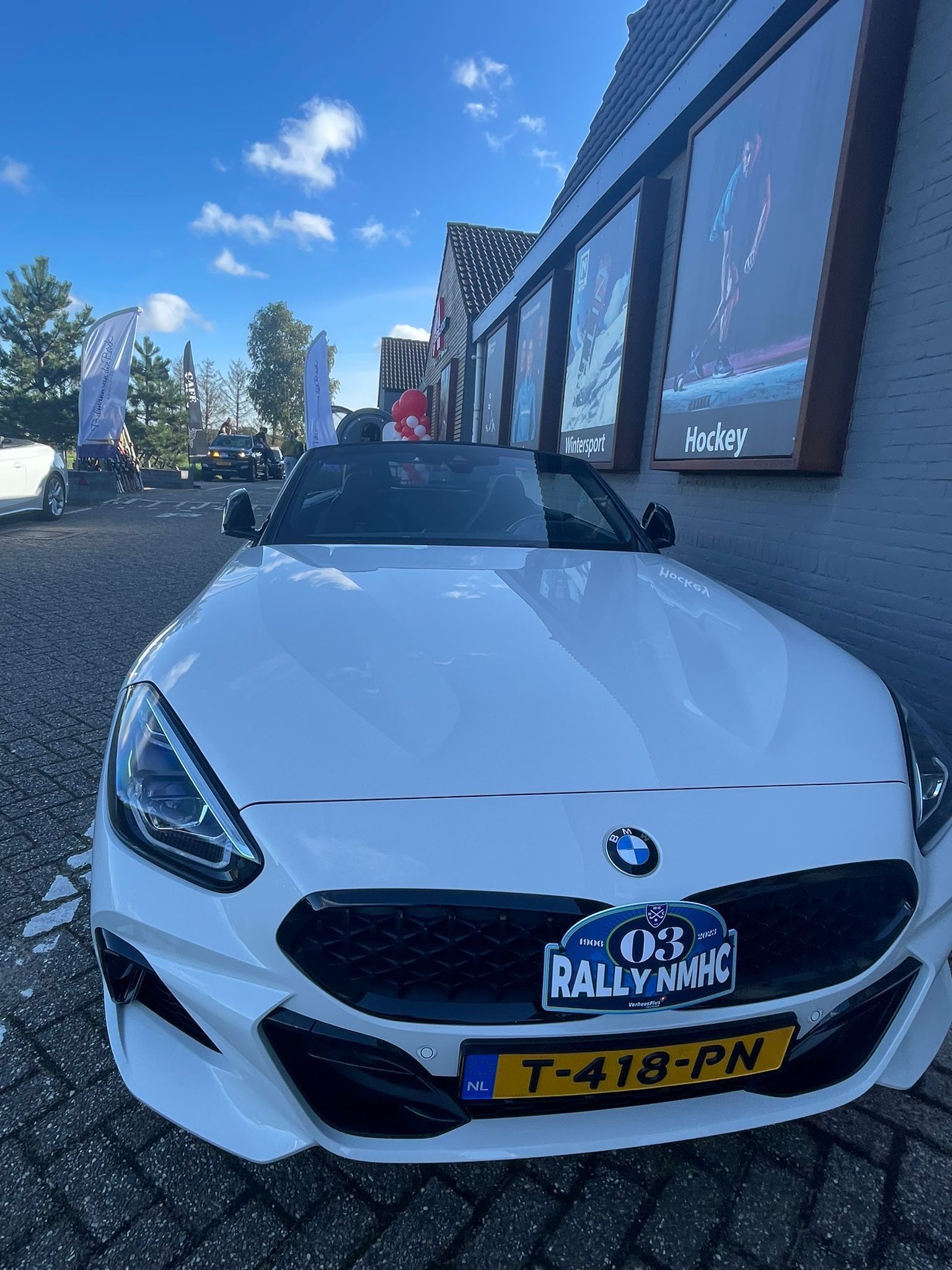 Een witte BMW Z4 roadster staat geparkeerd voor een gebouw; een rallysticker op de grille.
