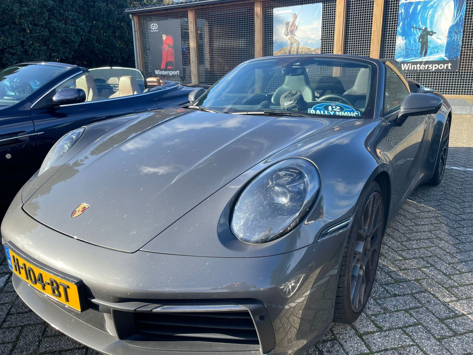 Een grijze Porsche 911 staat buiten geparkeerd, met links een cabriolet.