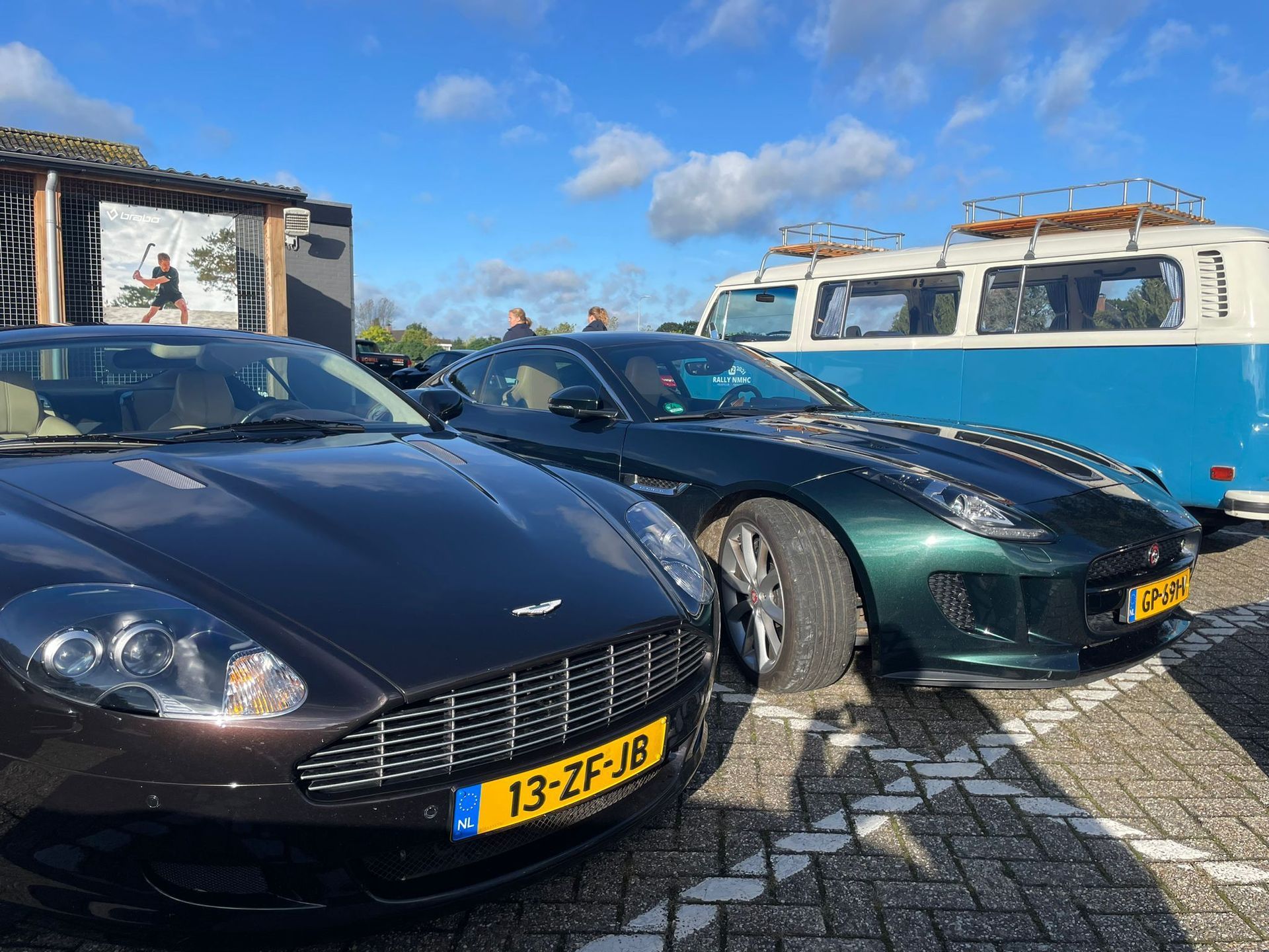 Donkere Aston Martins en groene Jaguars staan ​​geparkeerd naast een blauw-witte VW-bestelwagen op een zonnige dag.