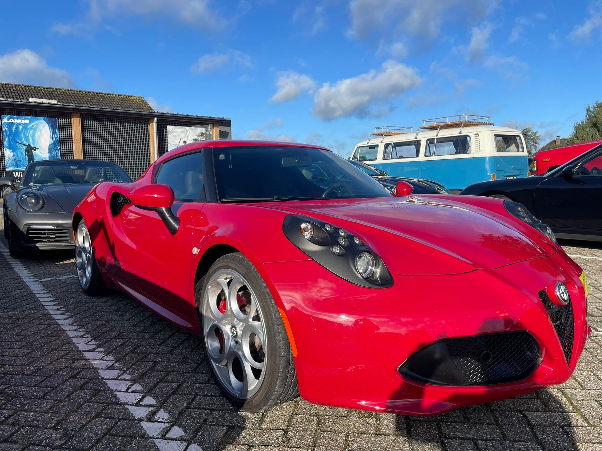 Een rode Alfa Romeo 4C sportwagen staat geparkeerd op een verharde parkeerplaats op een zonnige dag.